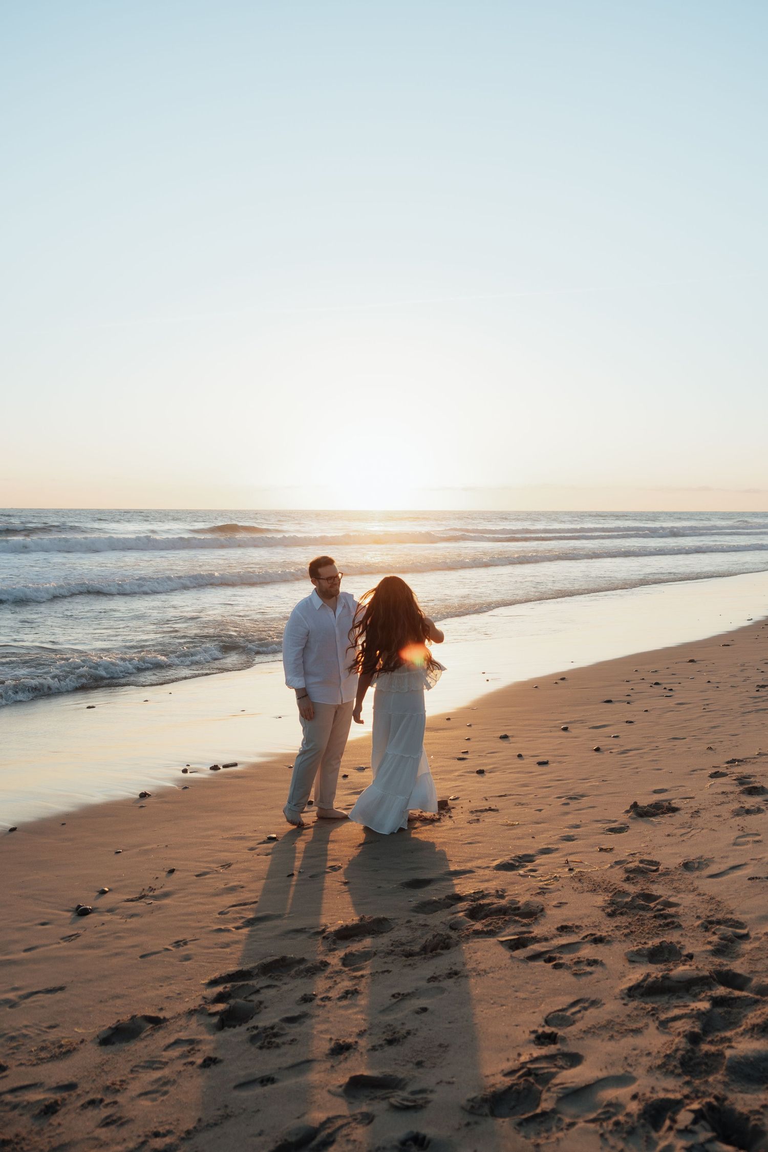 Couple in wedding attire walking hand in hand on a sandy beach at golden sunset with ocean waves.