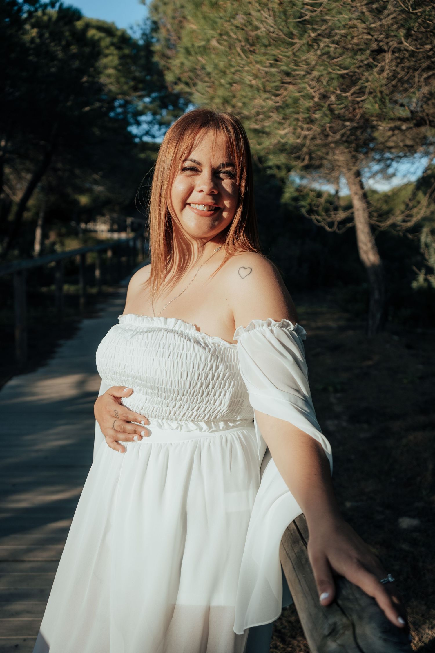 Smiling woman in white off-shoulder dress poses on a sunny tree-lined path.