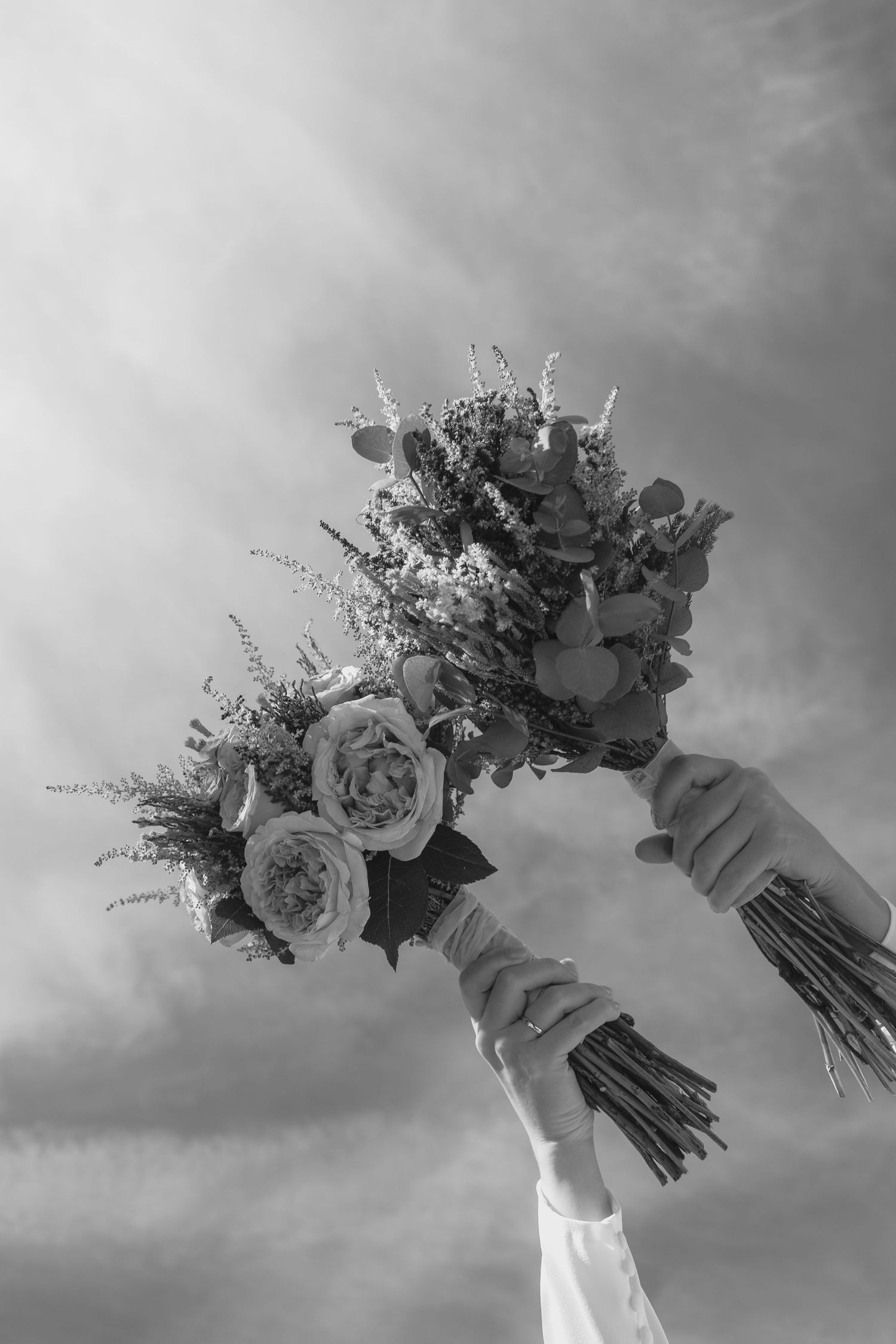 A bride raises a beautiful bouquet of roses and eucalyptus against a dramatic cloudy sky.