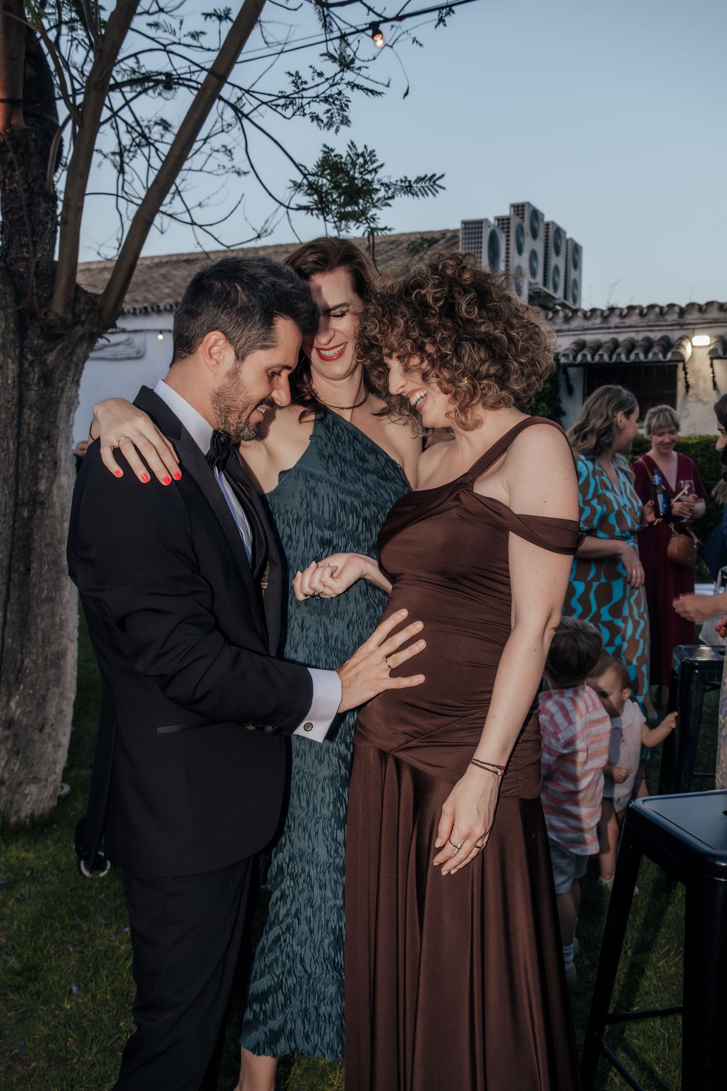 Pregnant woman in brown dress with friends at outdoor wedding reception celebration.