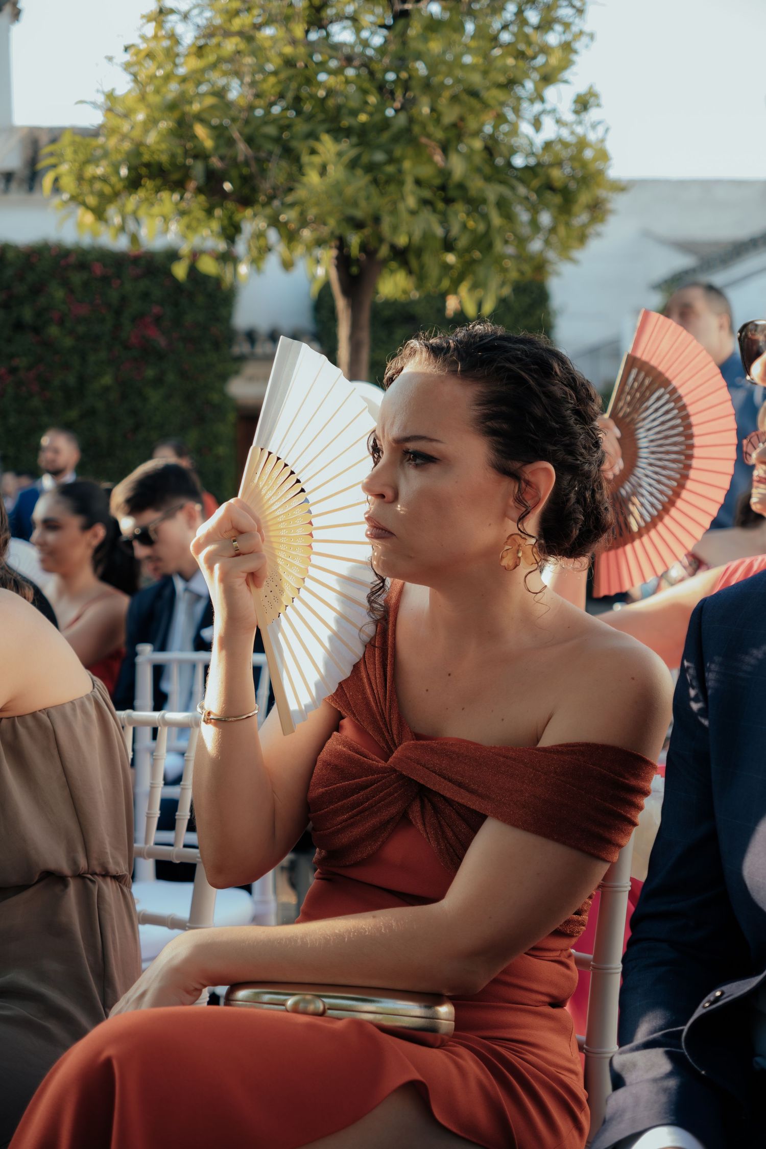 Elegant woman in rust off-shoulder dress fans herself with white hand fan at outdoor wedding.