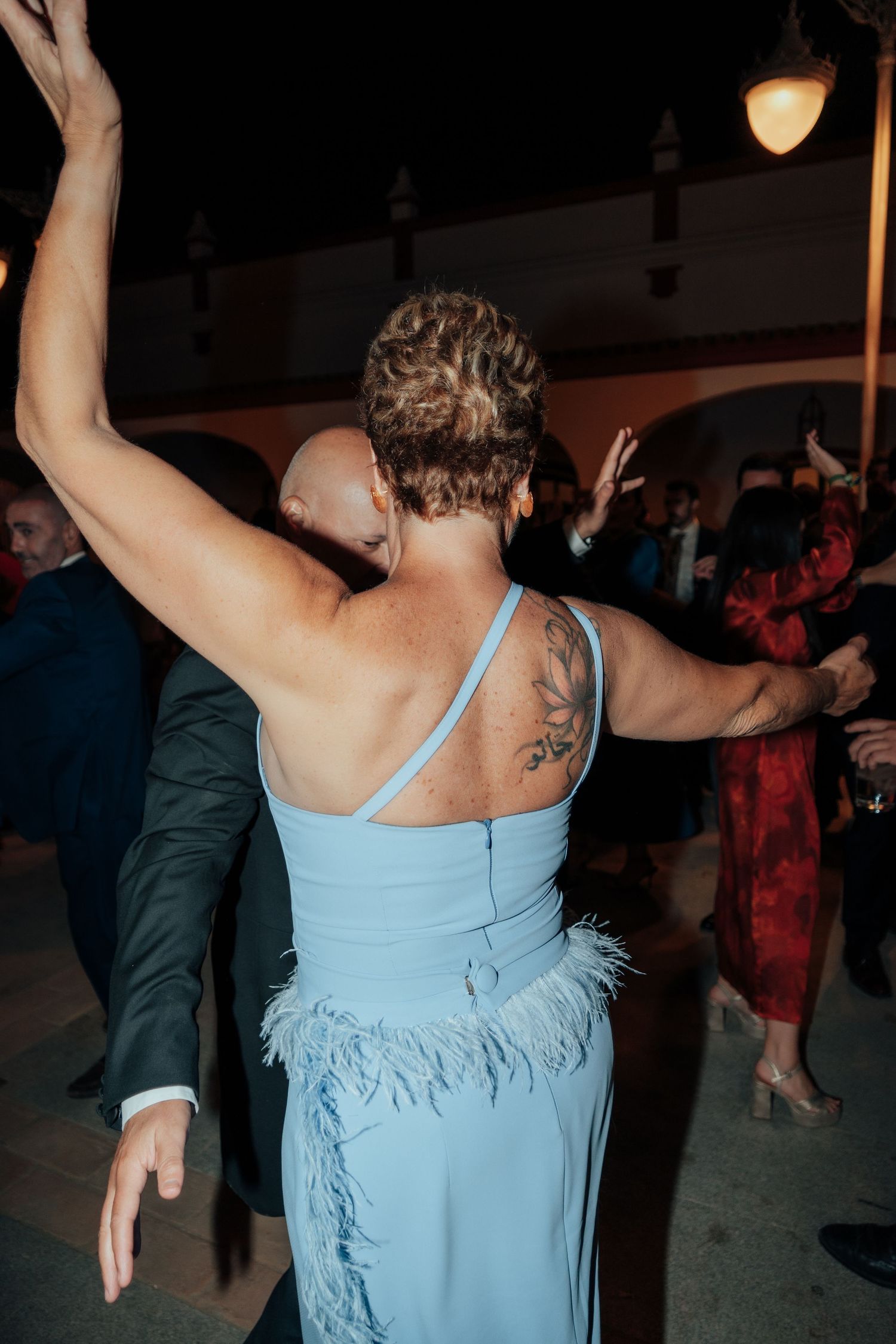Woman in blue feathered dress with floral tattoo dancing with arms raised at wedding.
