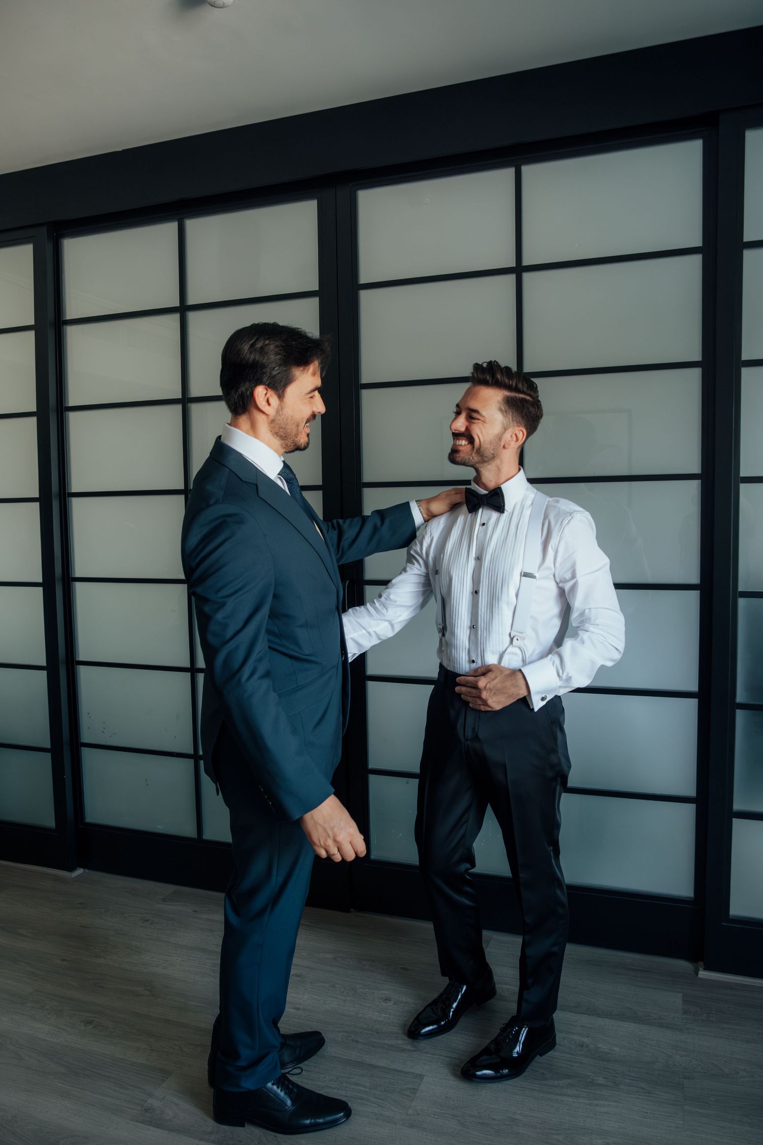 Two well-dressed men in suits share a smile while one adjusts the other's bow tie indoors.