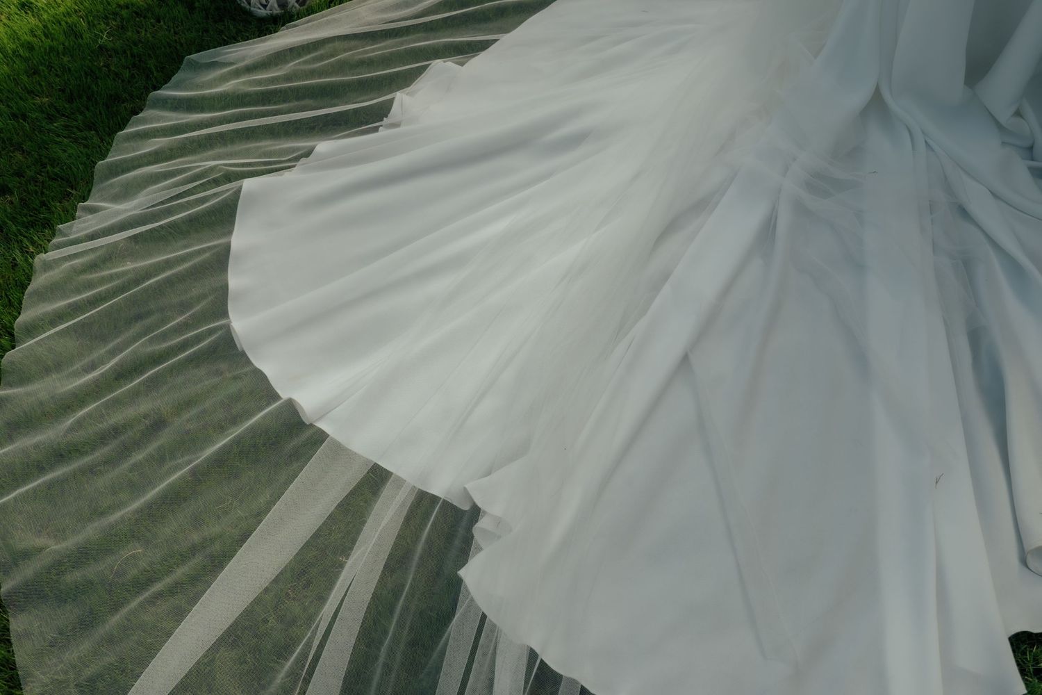 Close-up aerial view of a flowing white bridal veil and wedding dress train spread across green grass.