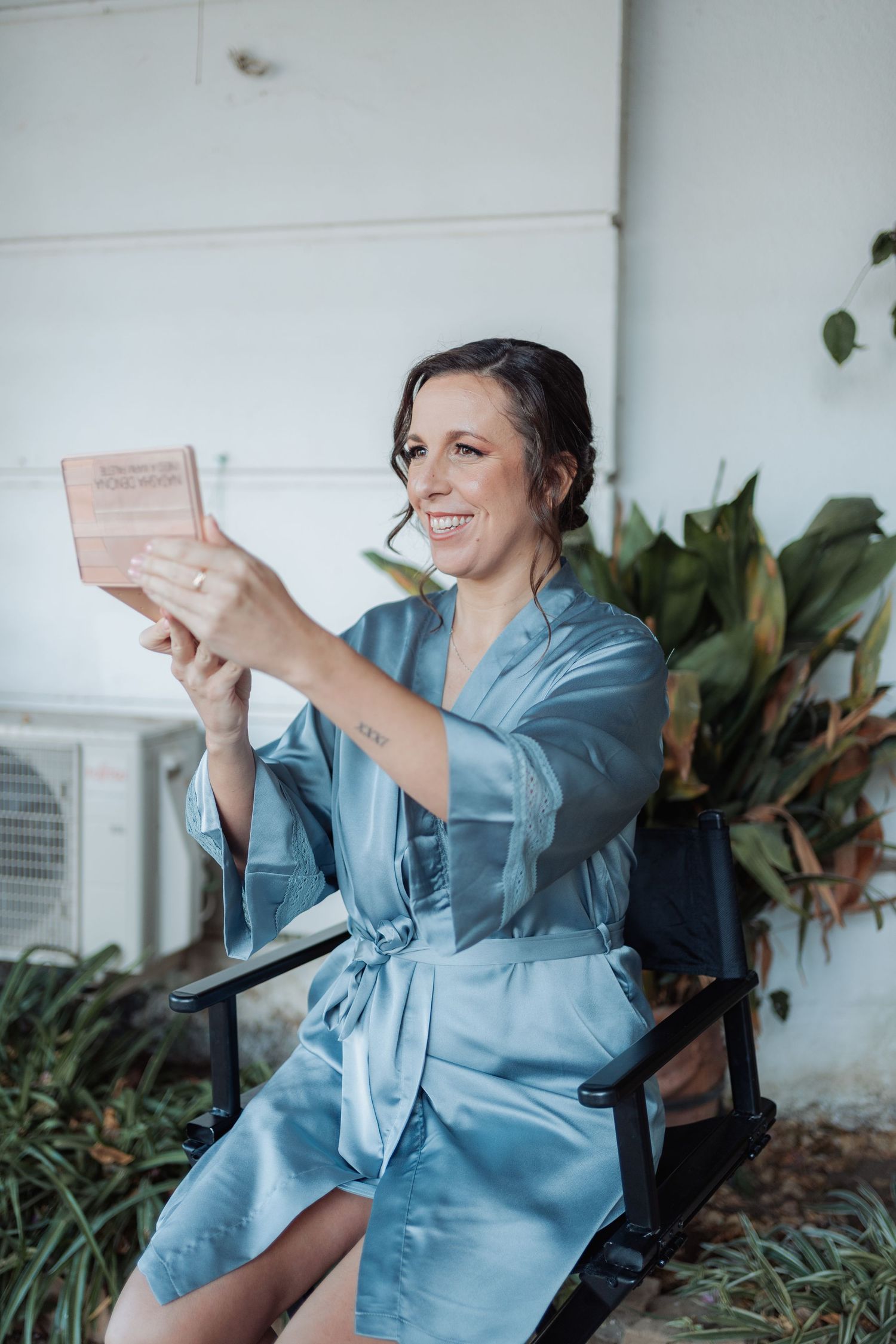 Smiling bride in blue satin robe reading wooden keepsake box while seated outdoors.