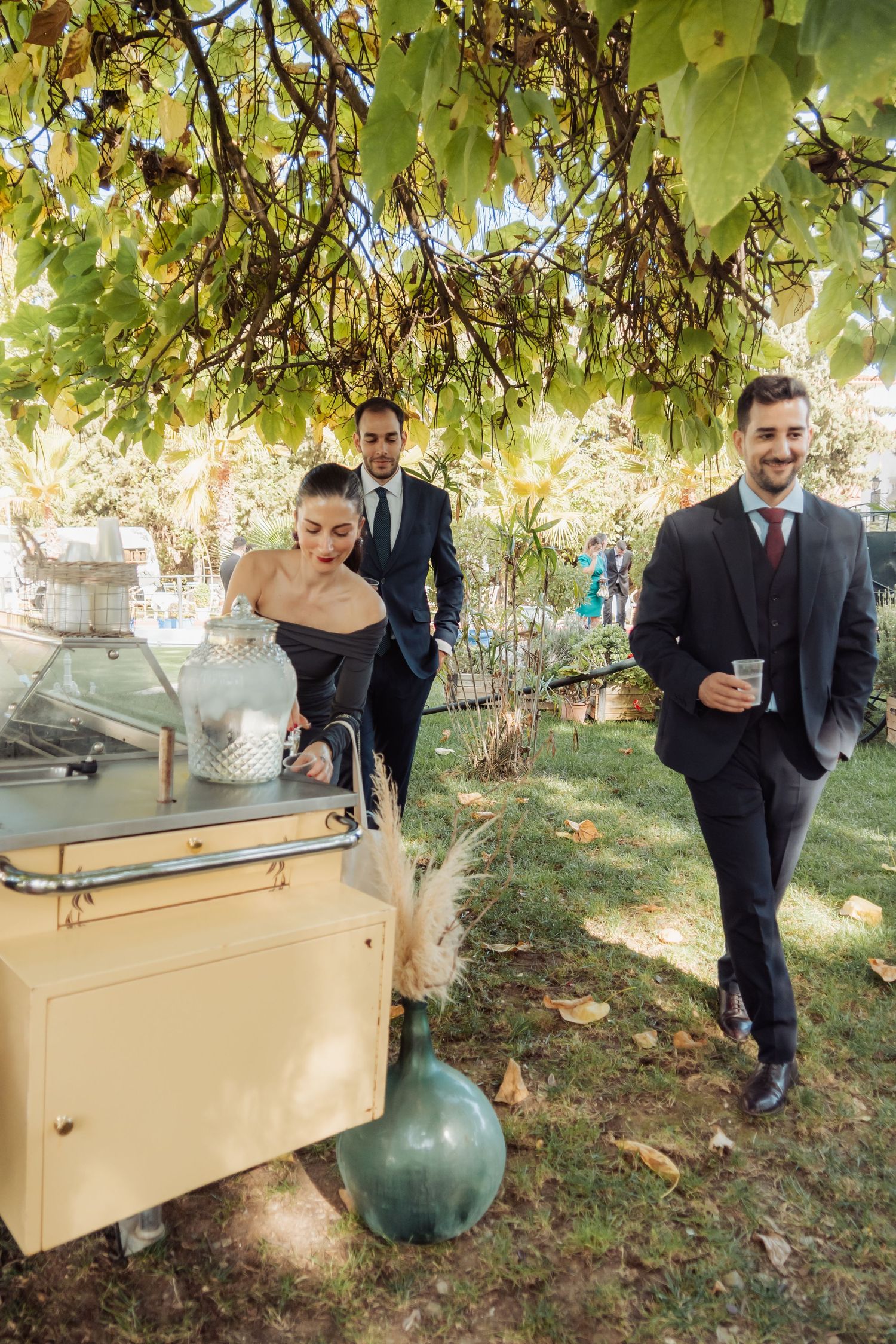 Smiling wedding guests in formal attire gather near a vintage beverage cart under leafy grapevine canopy.