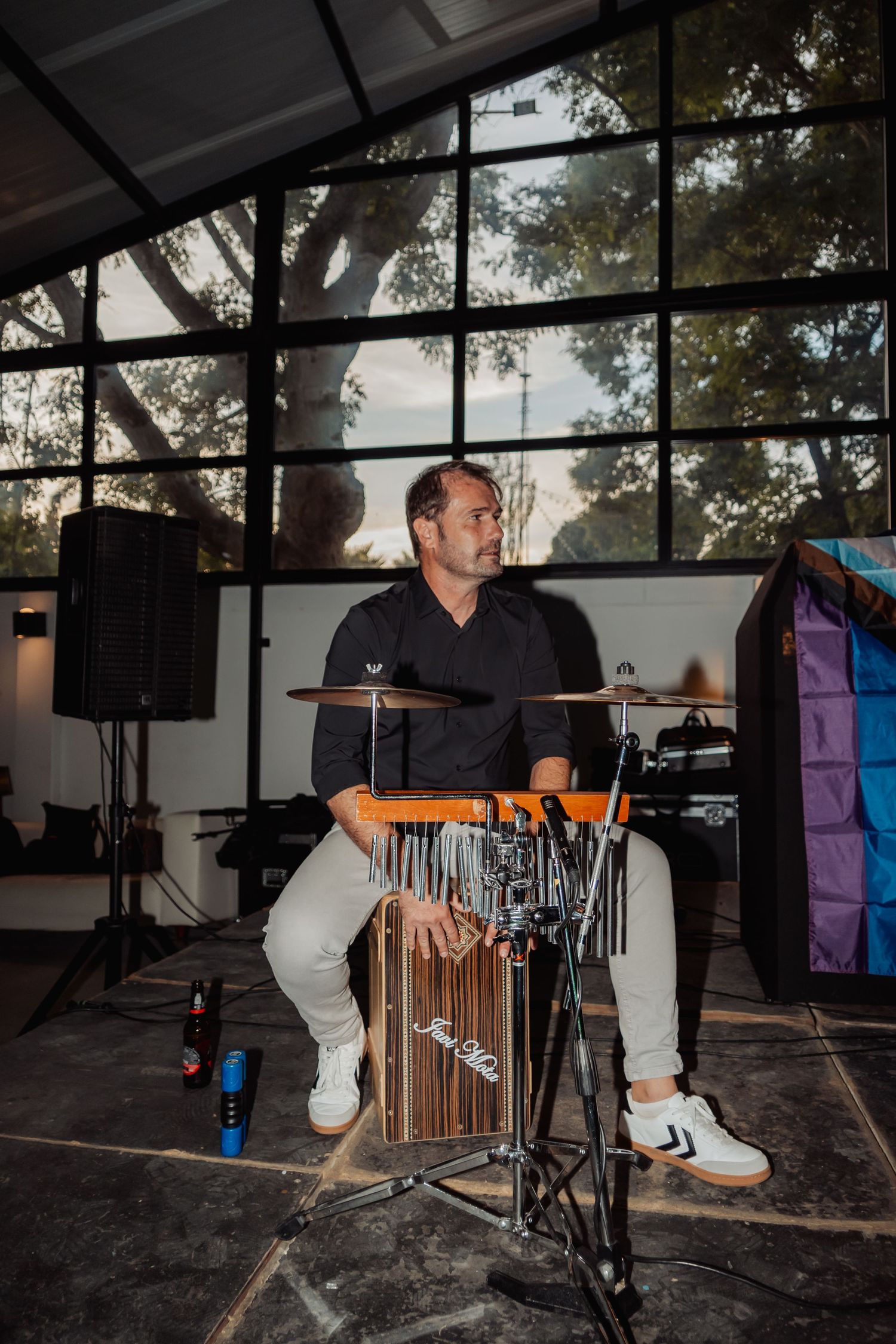 Male drummer performing on a cajon drum set in a glass-walled venue during a live event.