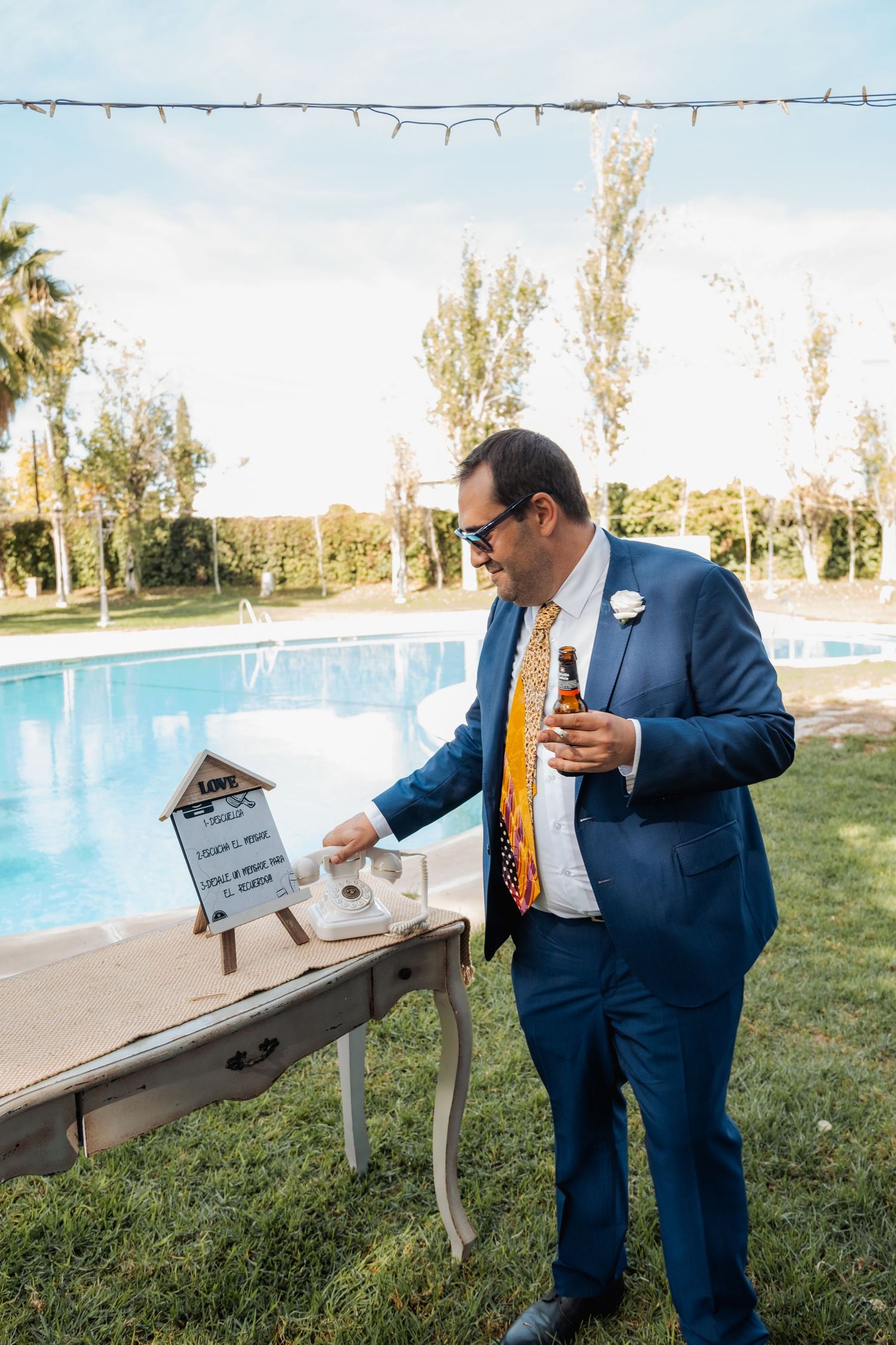 Groom in blue suit holding beer bottle at outdoor wedding by pool.