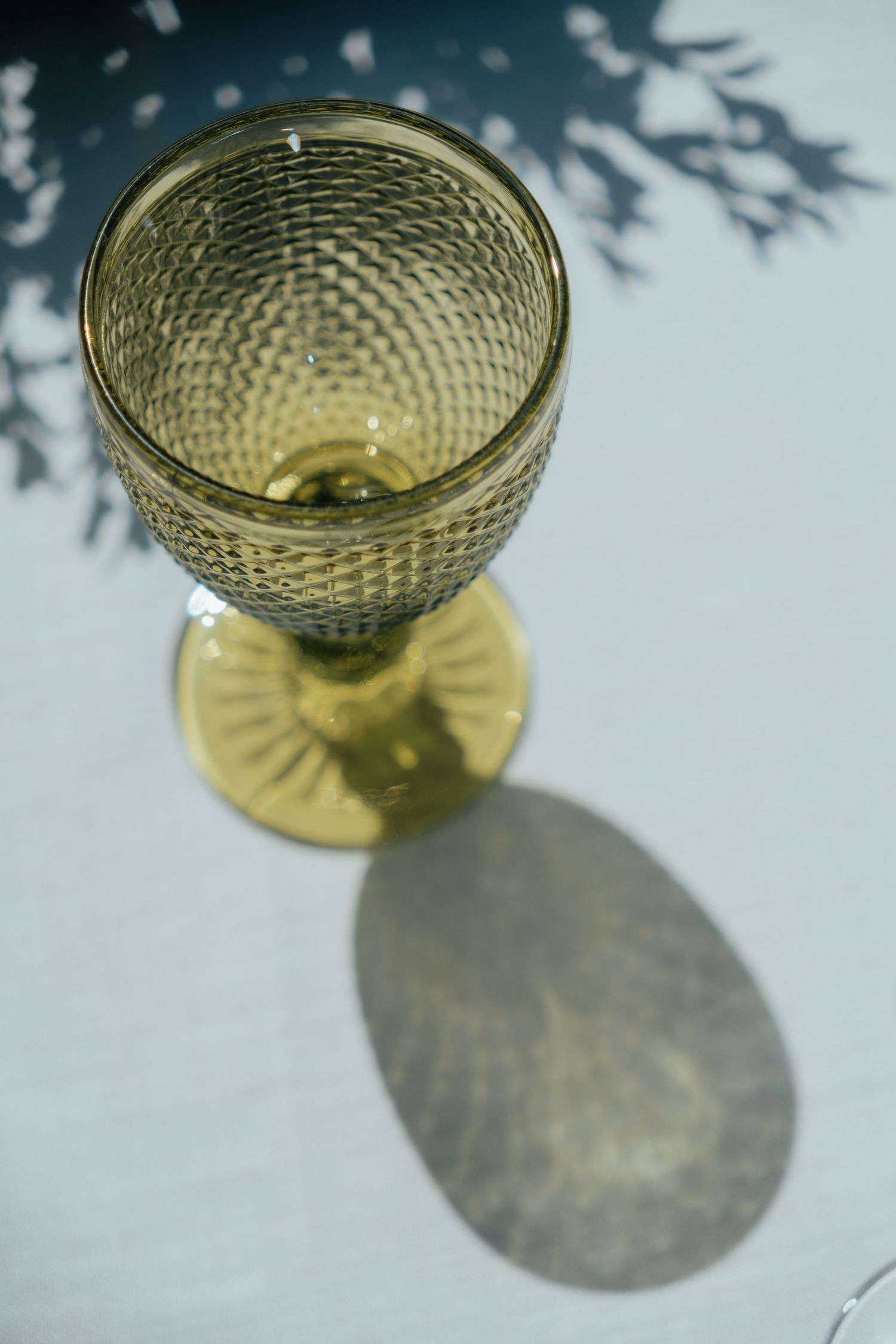 Elegant green textured glass goblet casting an oval shadow on a white linen tablecloth in natural sunlight.