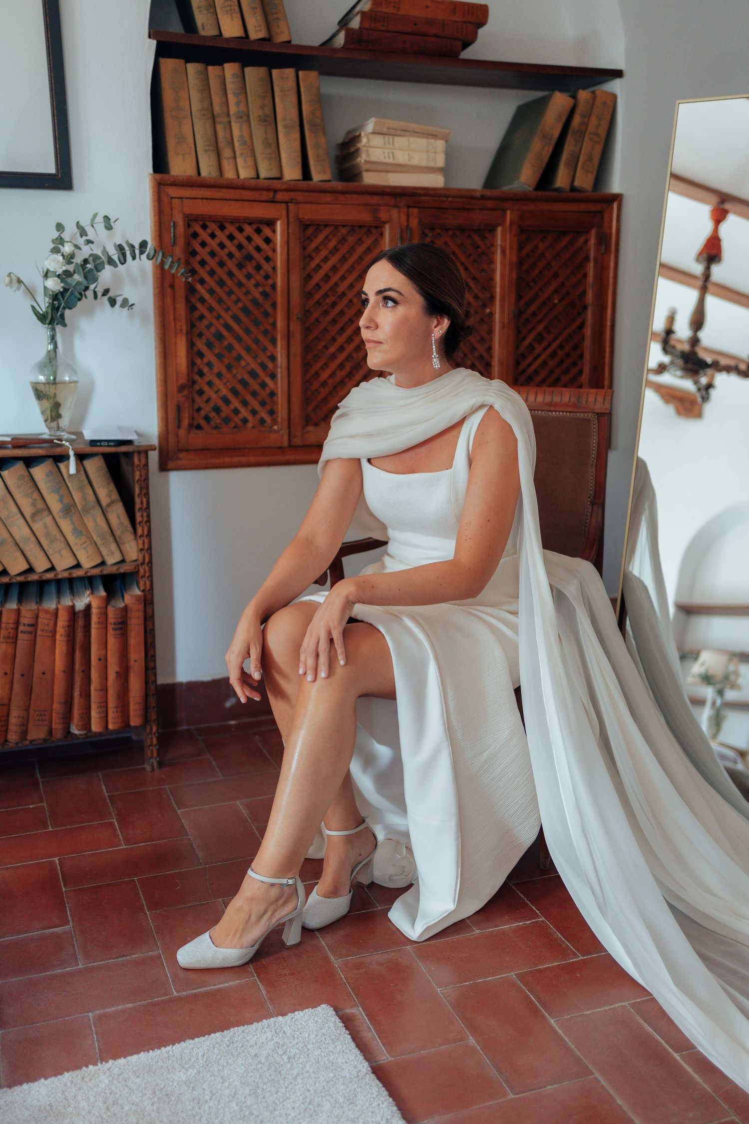 Bride in sleek white wedding gown with cape sitting elegantly beside vintage wooden bookcase.