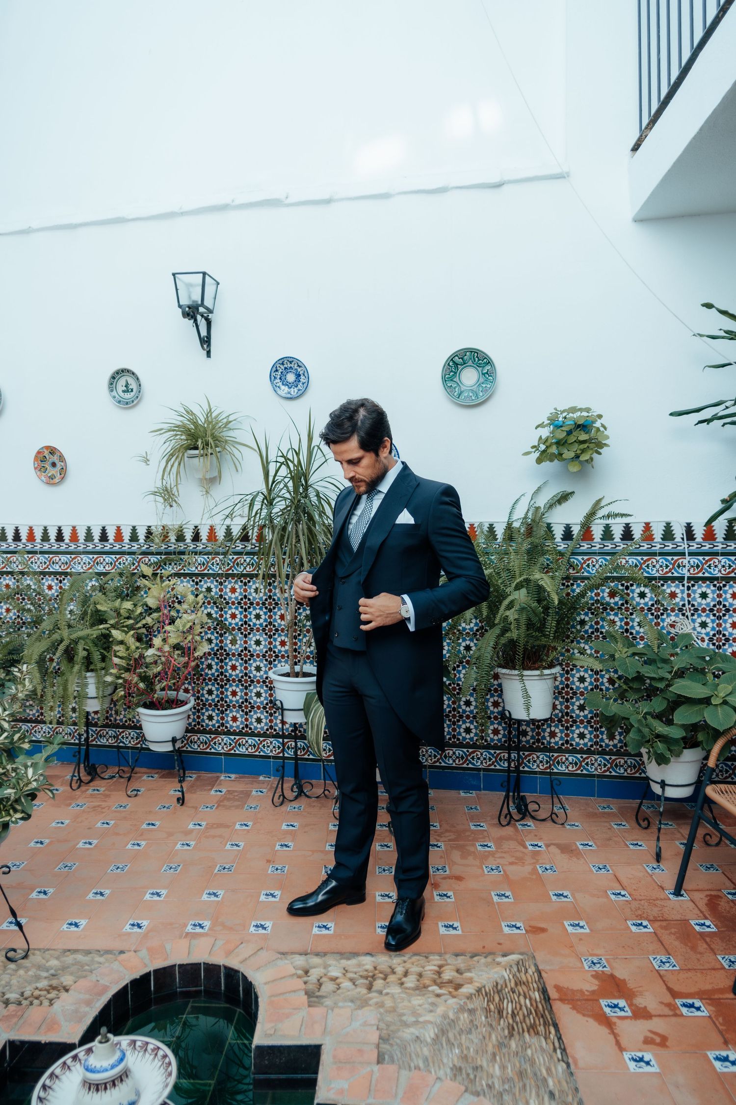Elegant groom in dark suit standing in Spanish courtyard with colorful mosaic tiles and potted plants.
