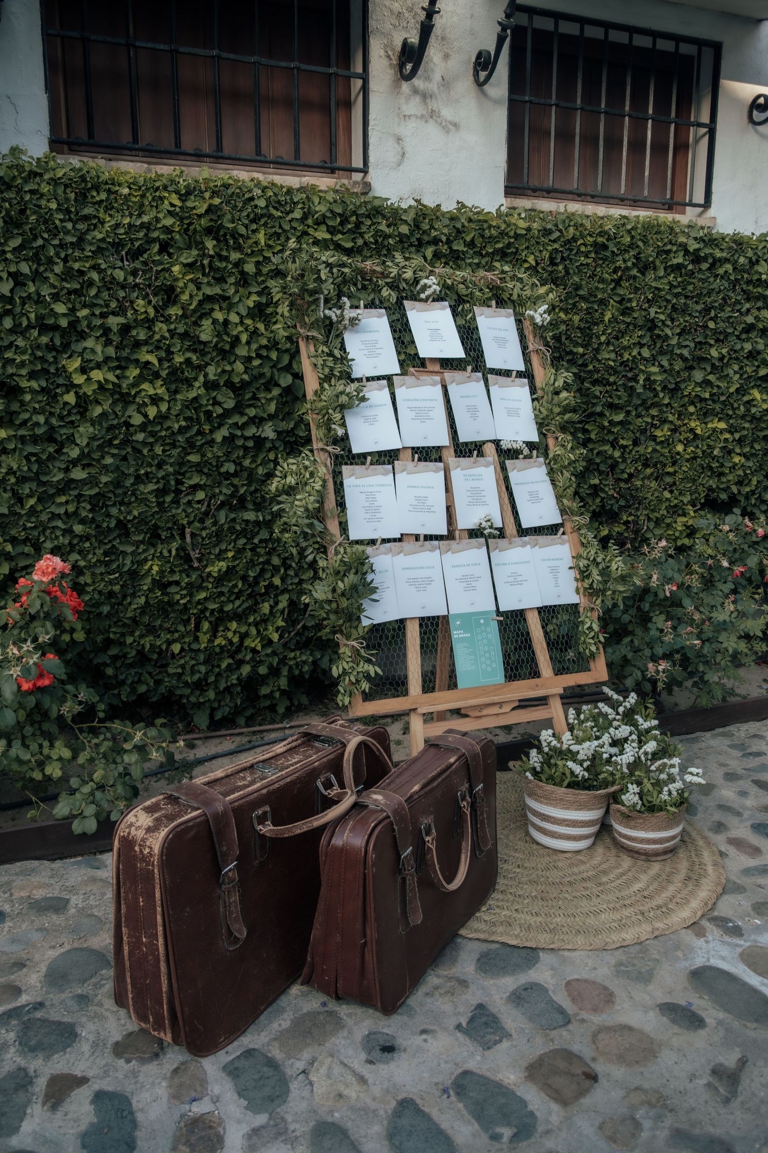Rustic wooden easel wedding seating chart display with greenery and vintage brown suitcases.