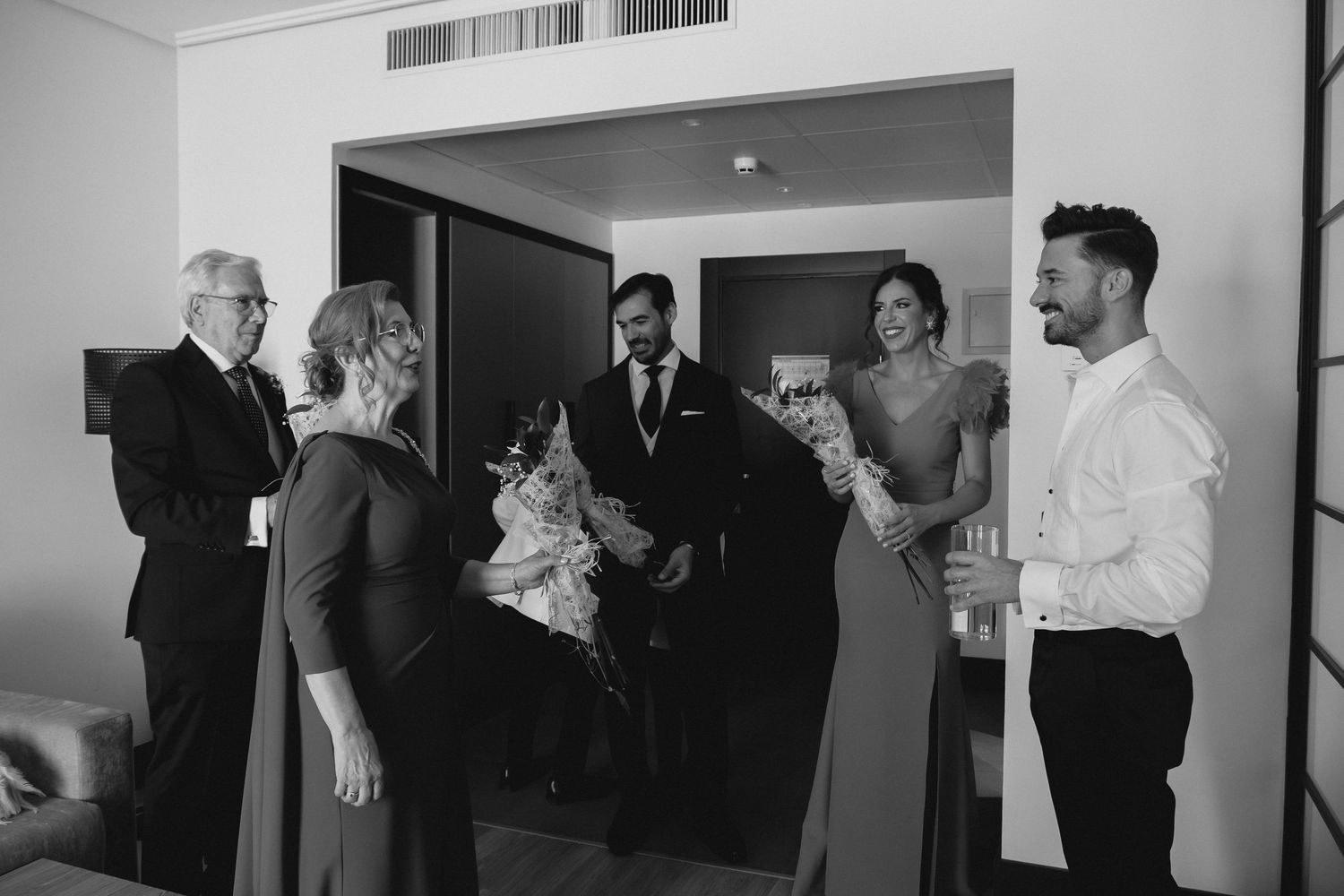Black and white photo of wedding guests greeting the bride and groom in a hotel room doorway.