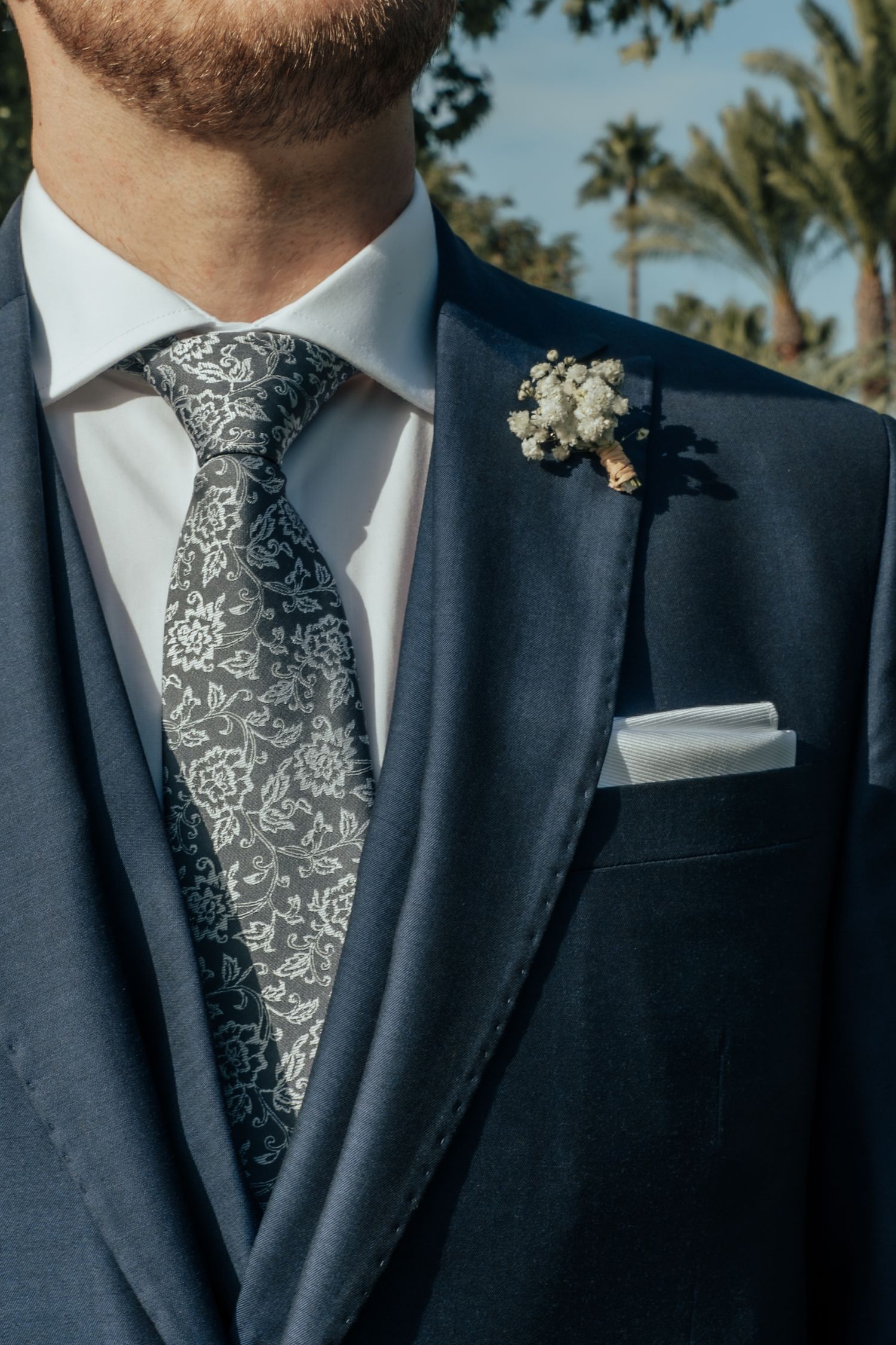 Close-up of groom in navy suit with floral tie, white pocket square, and baby's breath boutonniere.