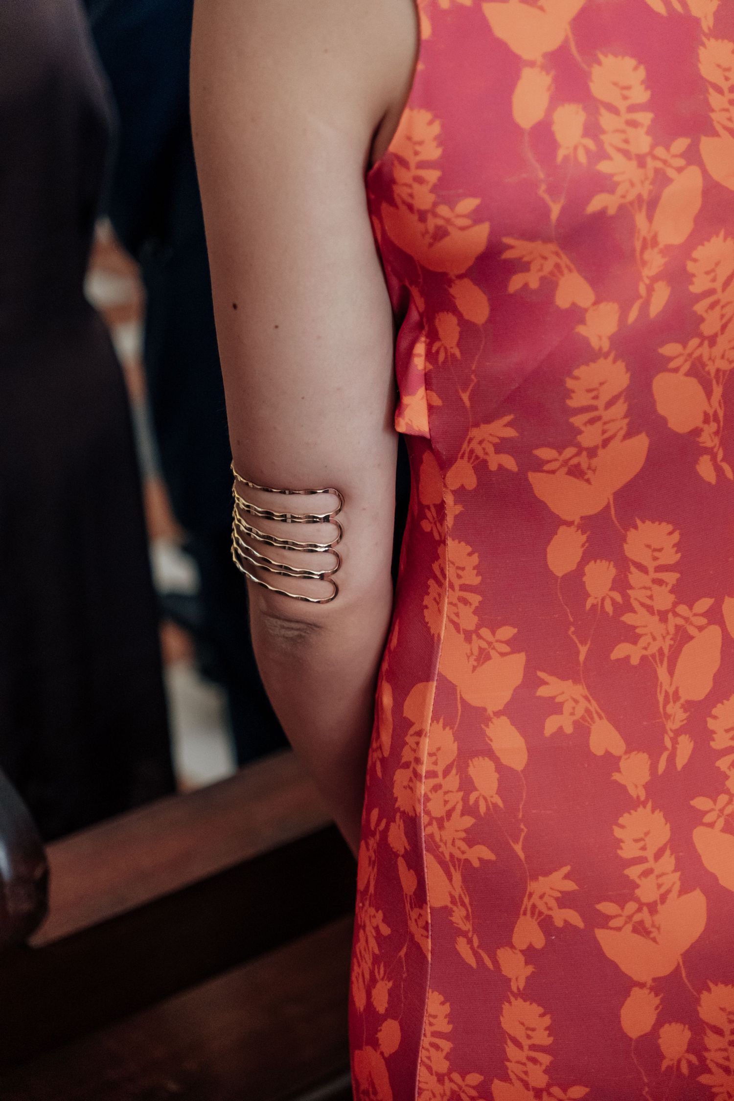 Close-up of woman wearing gold multi-band upper arm bracelet cuff with red floral print dress at wedding.