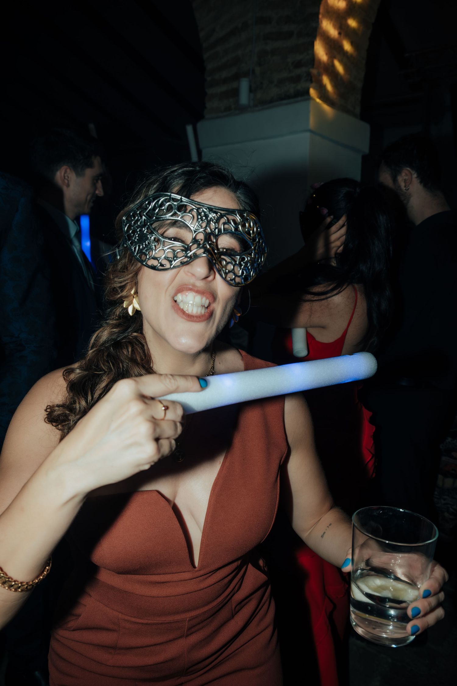 Woman in red dress wearing leopard masquerade mask holding glowing stick and drink at wedding party.