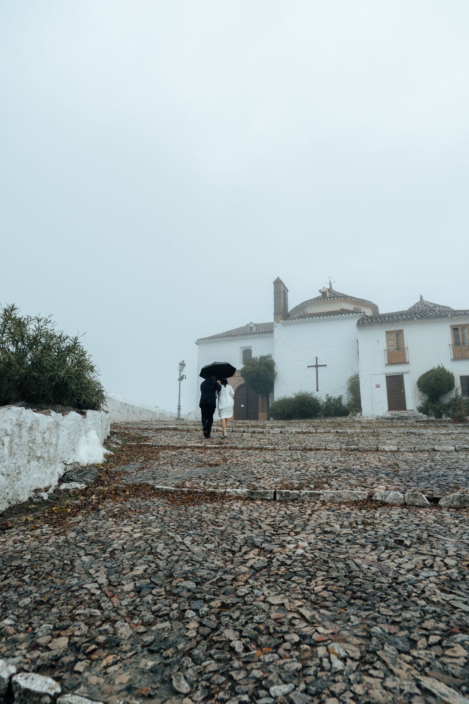 Wedding couple walking up cobblestone steps in fog toward white Spanish church with cross.