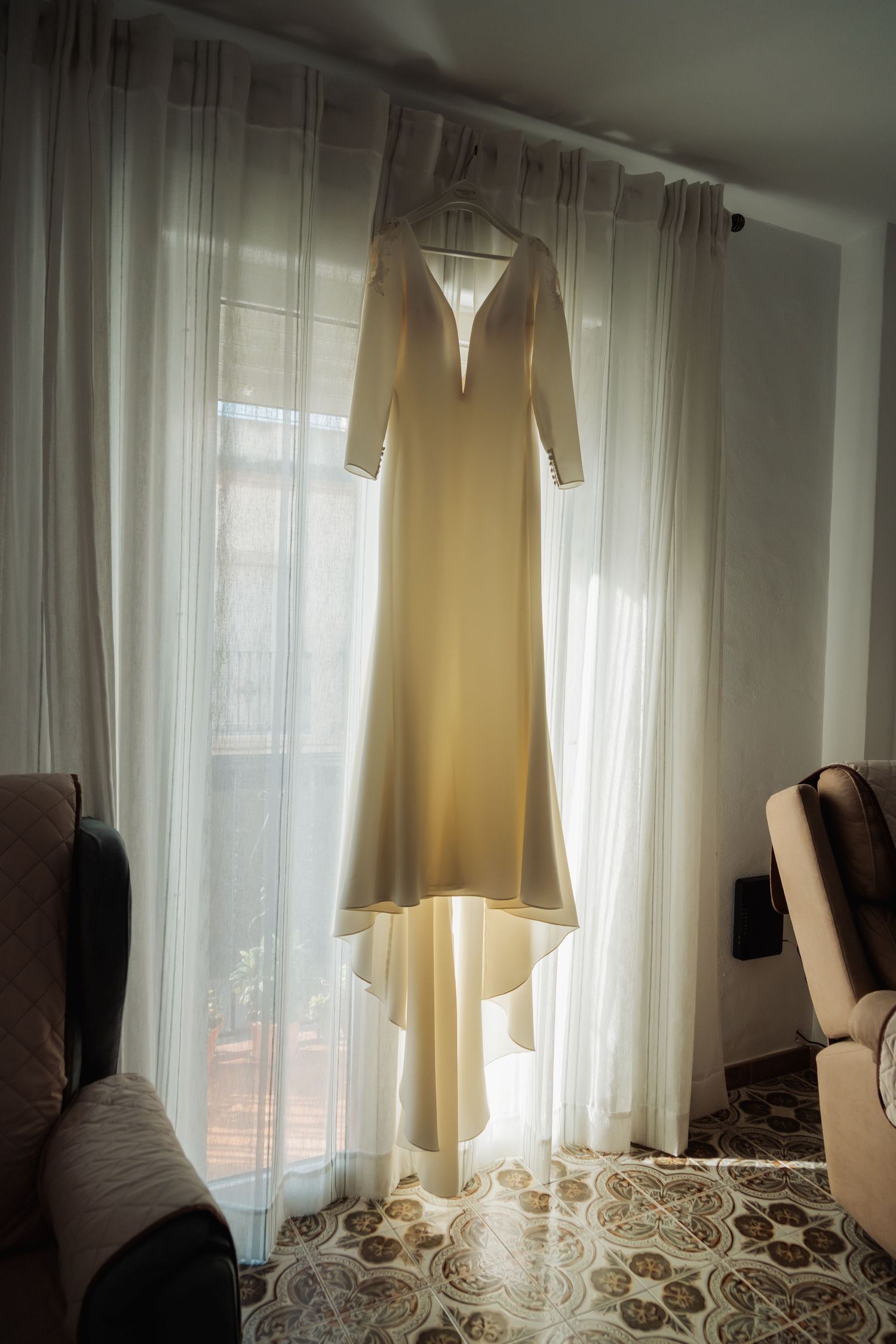 Elegant ivory long-sleeve wedding dress hanging on curtain rod in sunlit room.