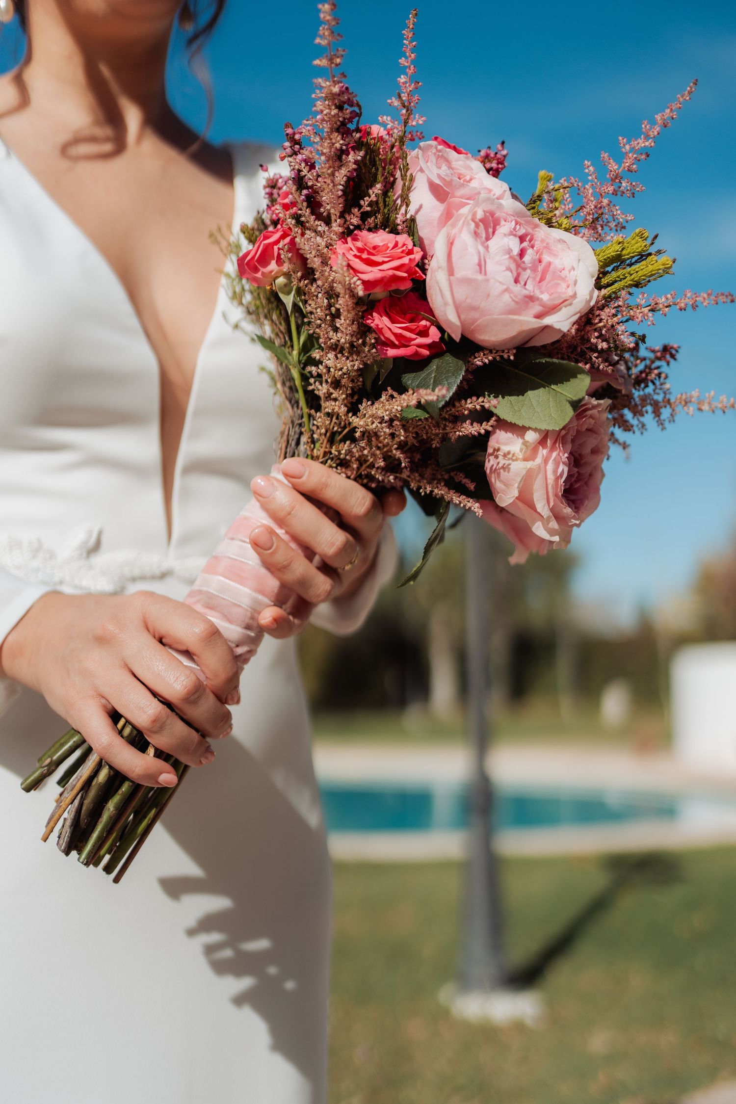 Bride holding lush pink peony and rose bridal bouquet with ribbon wrap outdoors.