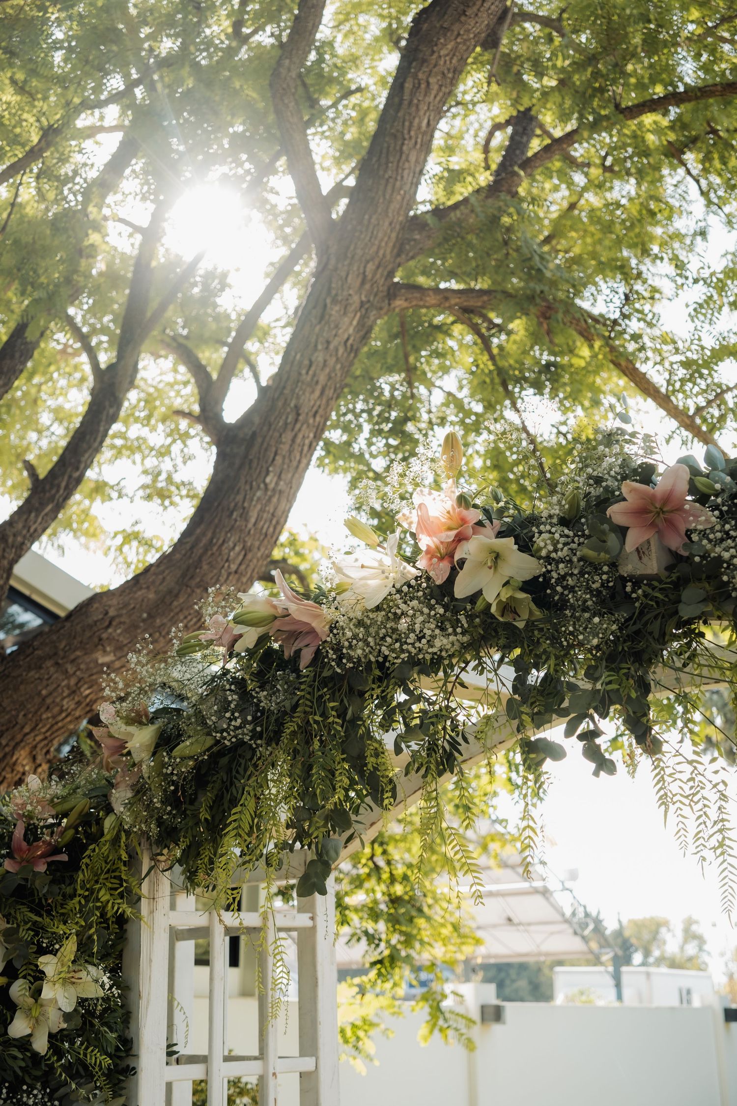 Floral arch with white lilies and pink blooms adorns a wedding ceremony altar beneath a sunlit tree.