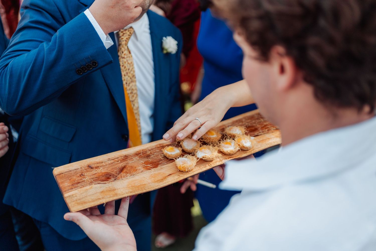 Wedding guest reaches for small appetizers on rustic wooden serving board at outdoor reception.