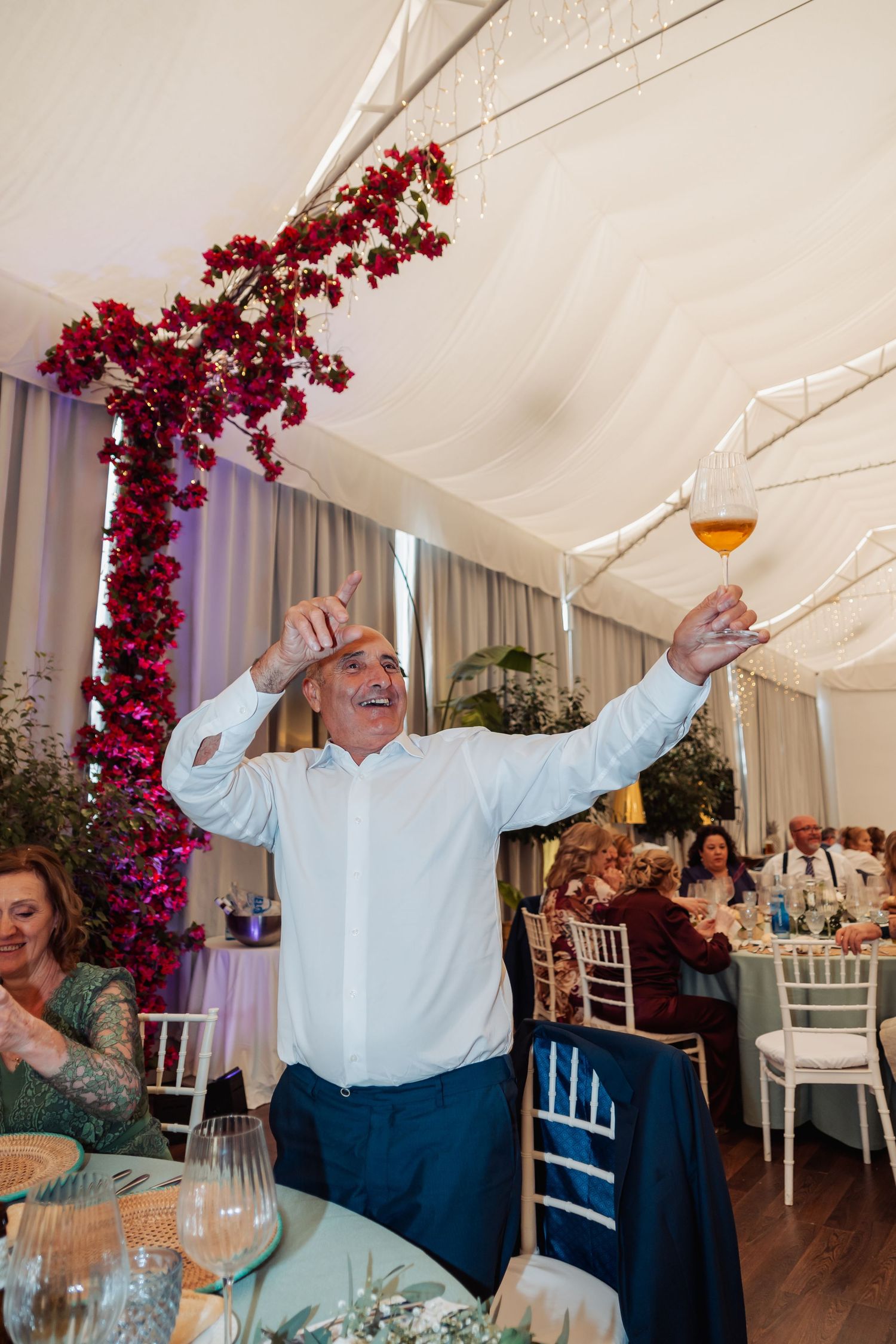 Cheerful older man raising a glass of wine and giving thumbs up at an elegant wedding reception.