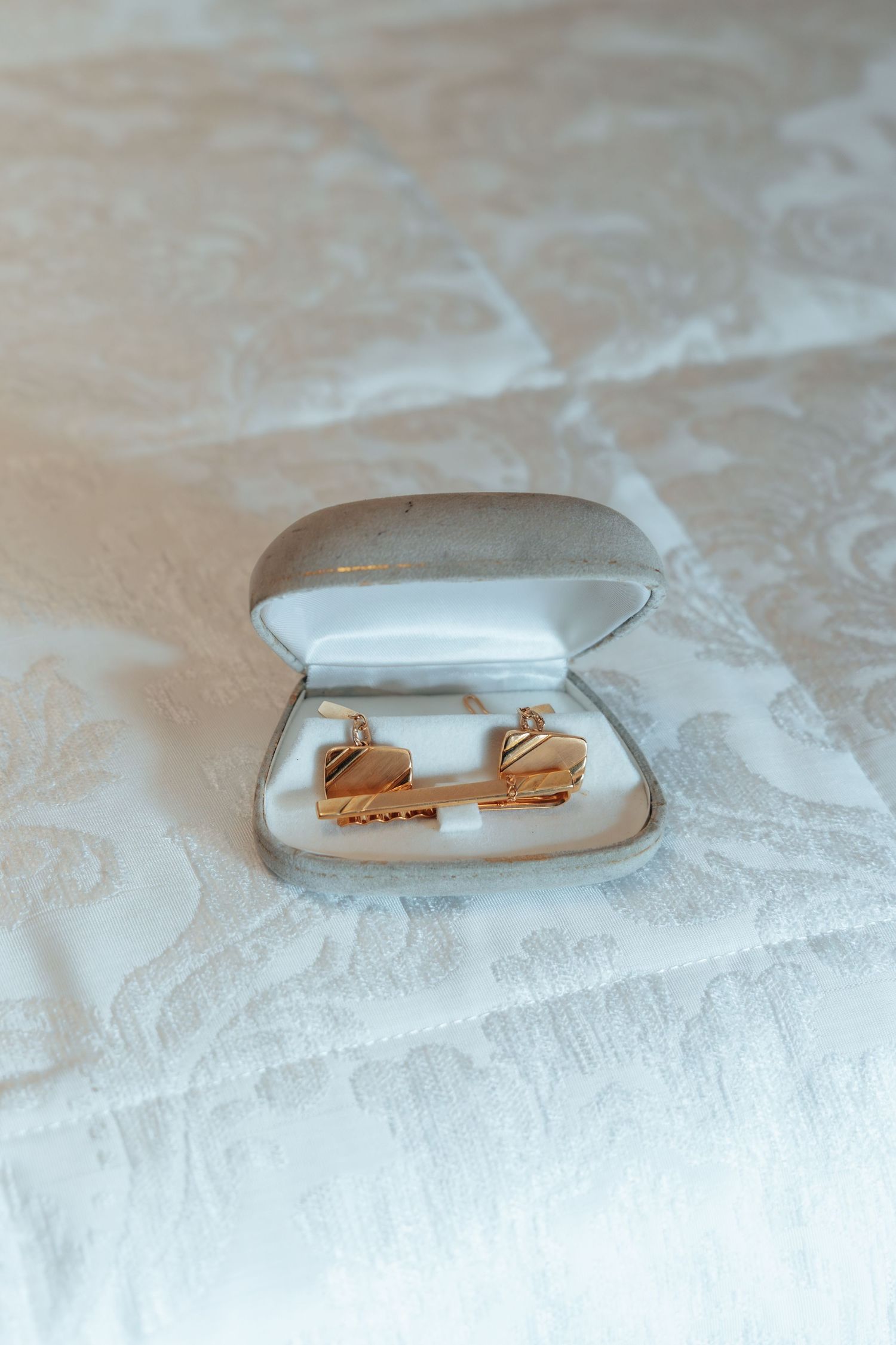 Gold cufflinks displayed in white jewelry box on embroidered white fabric for groom preparation.