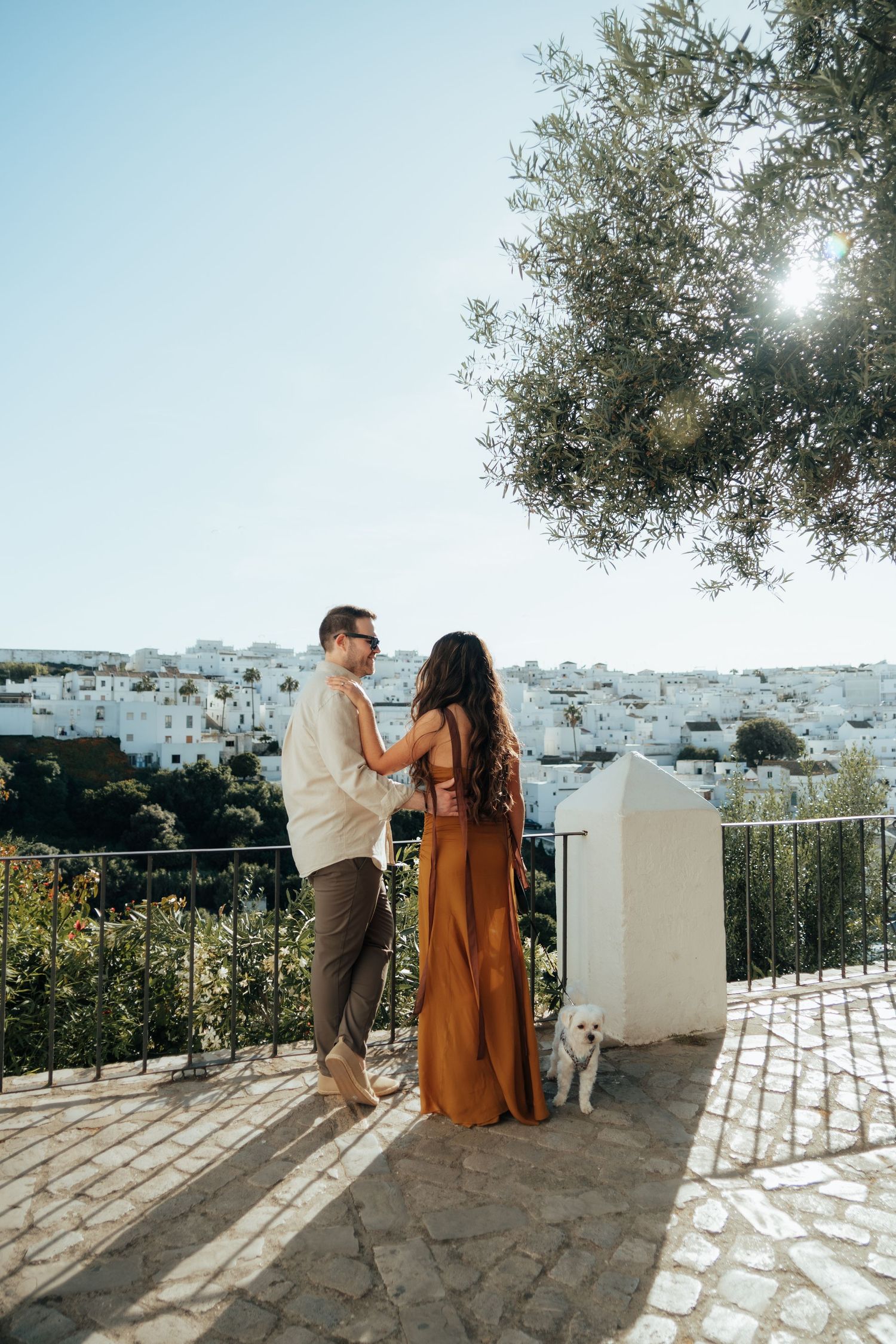Couple in elegant attire standing on a terrace with white village views, olive tree, and small dog in golden sunlight.