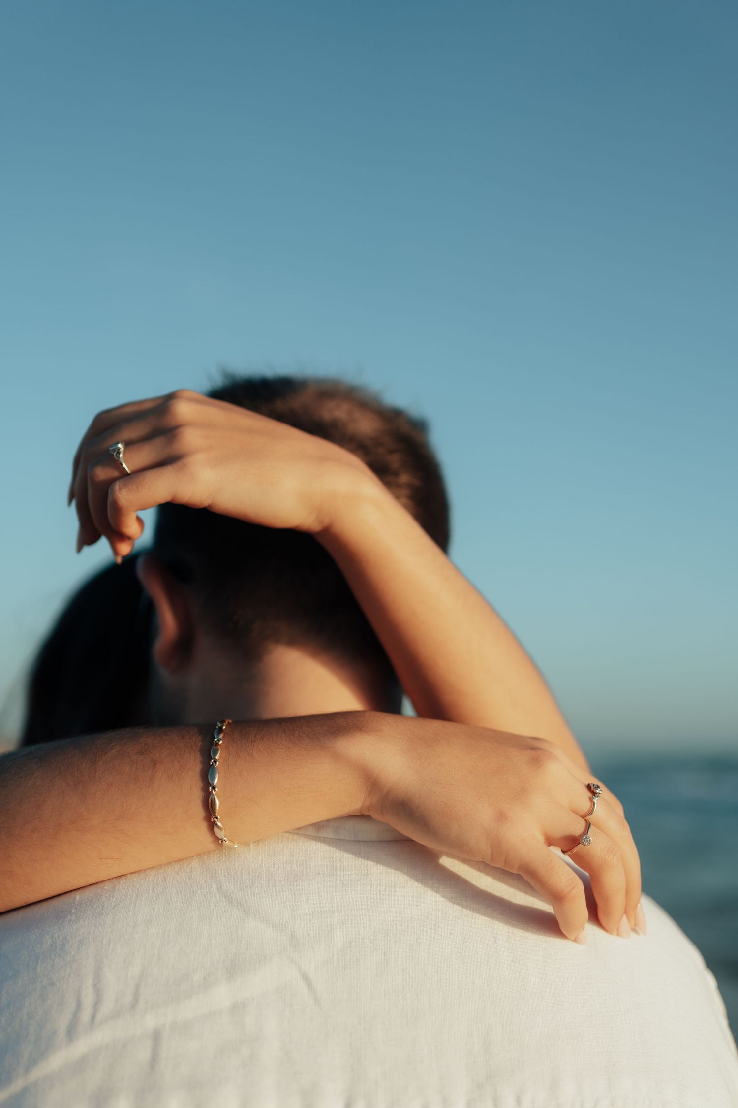 A couple embraces by the sea, woman's arms around man's neck, wearing gold rings and bracelet against blue sky.
