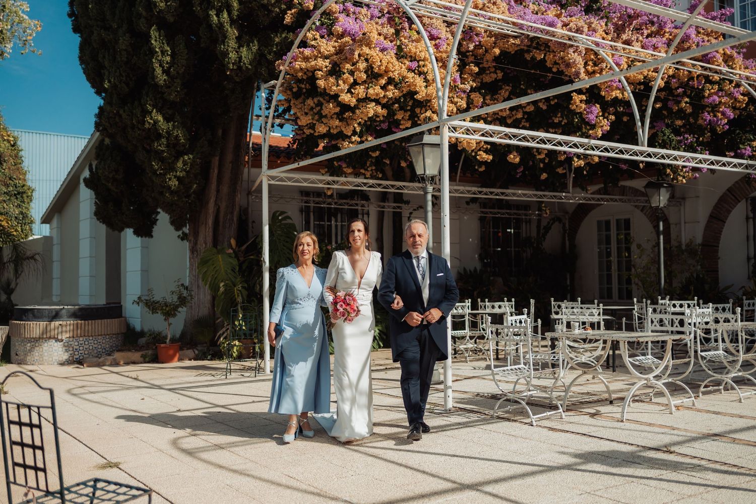 Bride and parents walk together under a white pergola with purple flowers at a sunny venue.