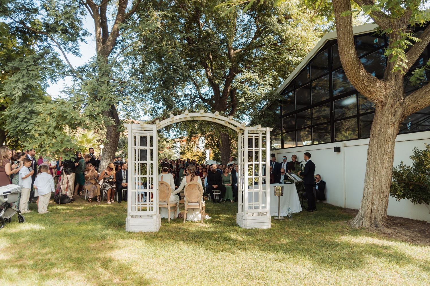 Outdoor wedding ceremony with white arch doors, seated guests, and glass building backdrop.