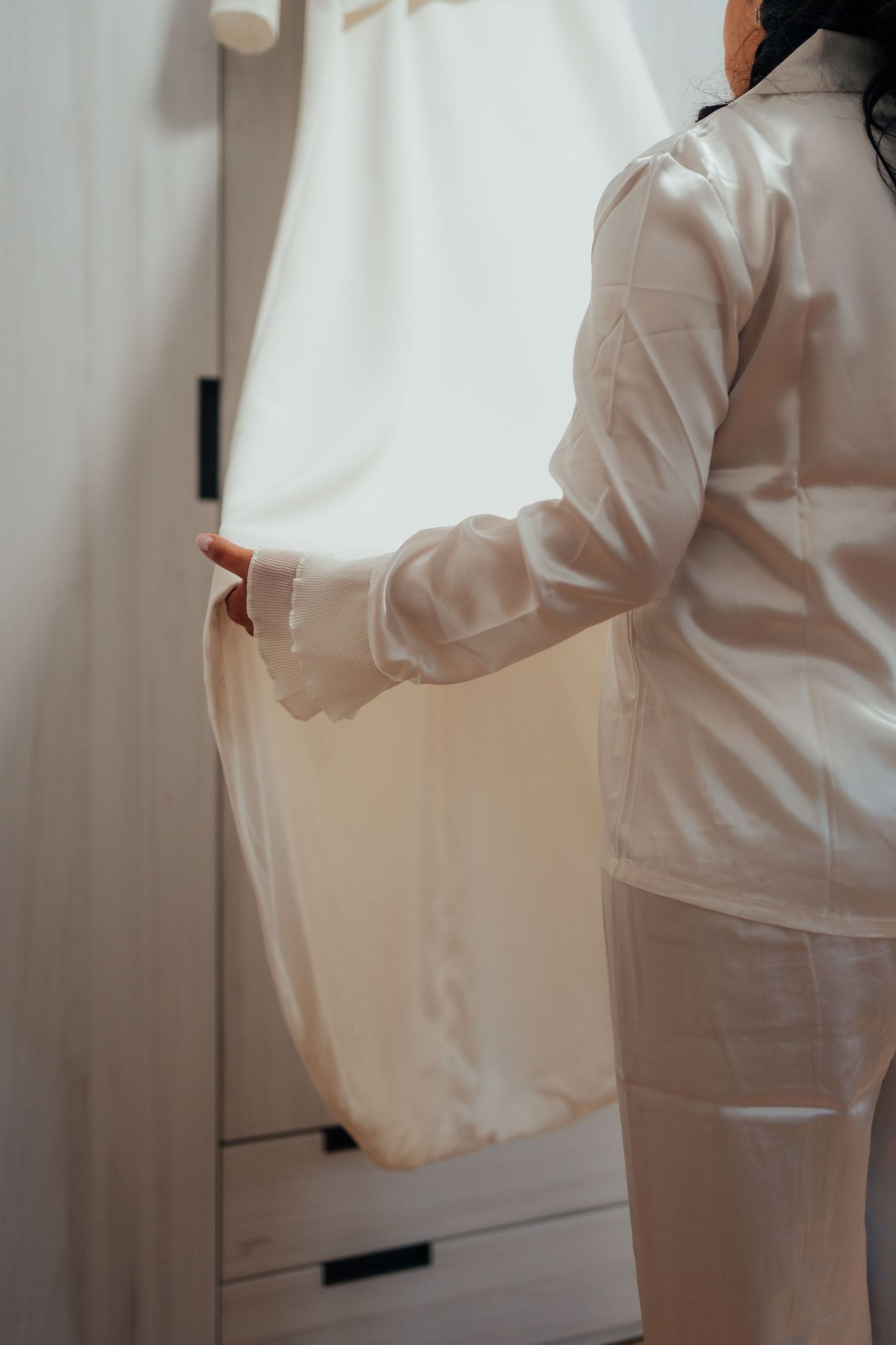 Bride in white satin robe holding wedding dress near wardrobe by bright window on wedding morning.