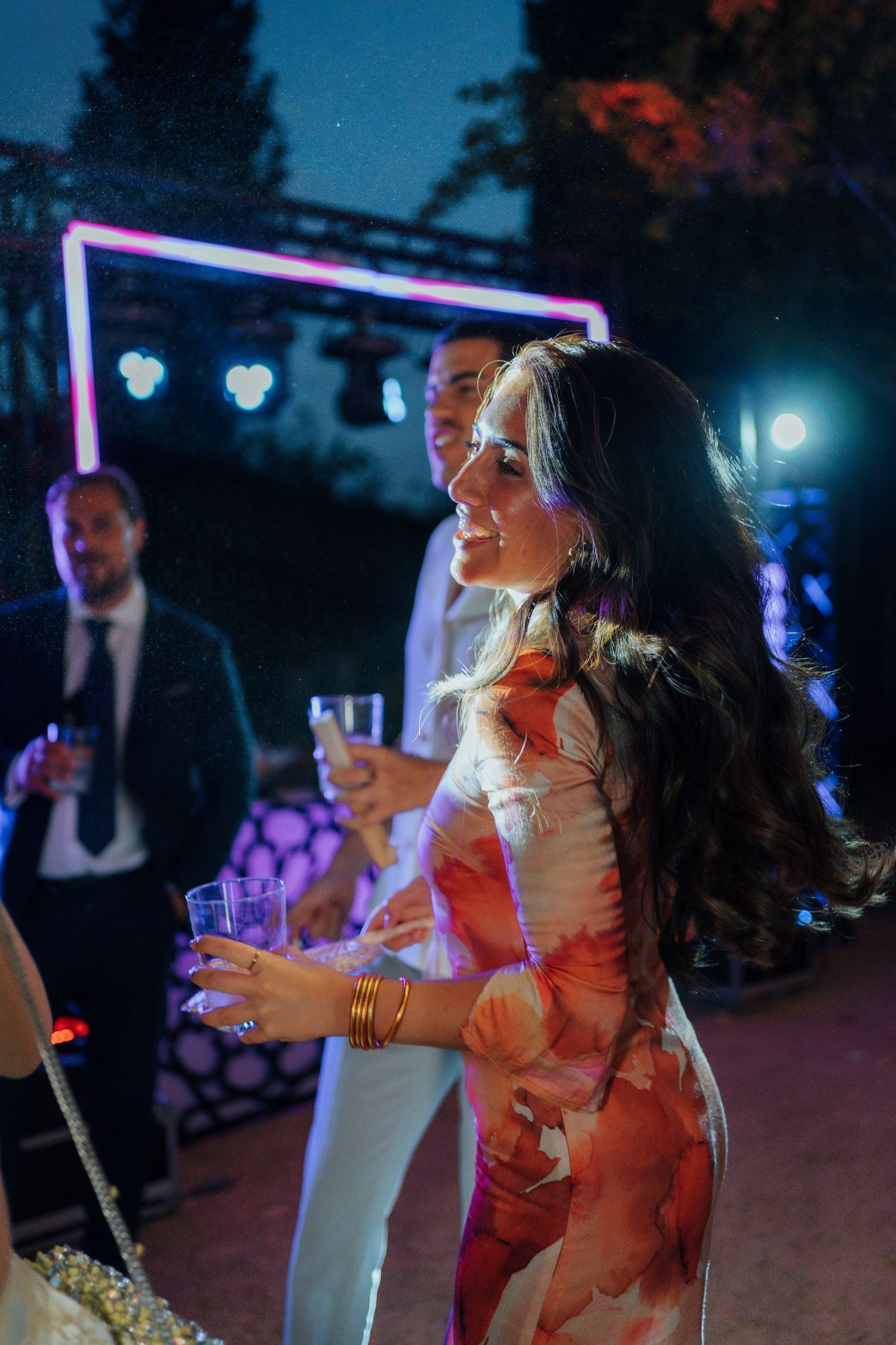 Smiling wedding guest in floral dress holds champagne glass at vibrant outdoor night reception.