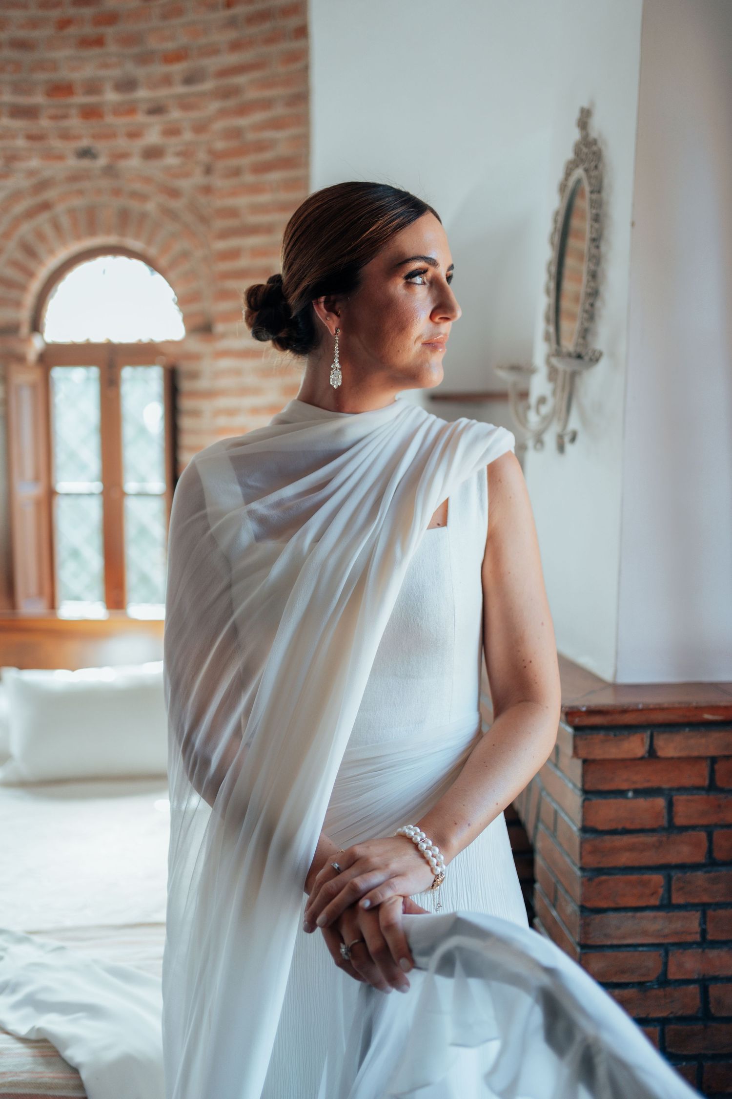 Bride in white draped wedding gown gazing out window in rustic brick interior room.