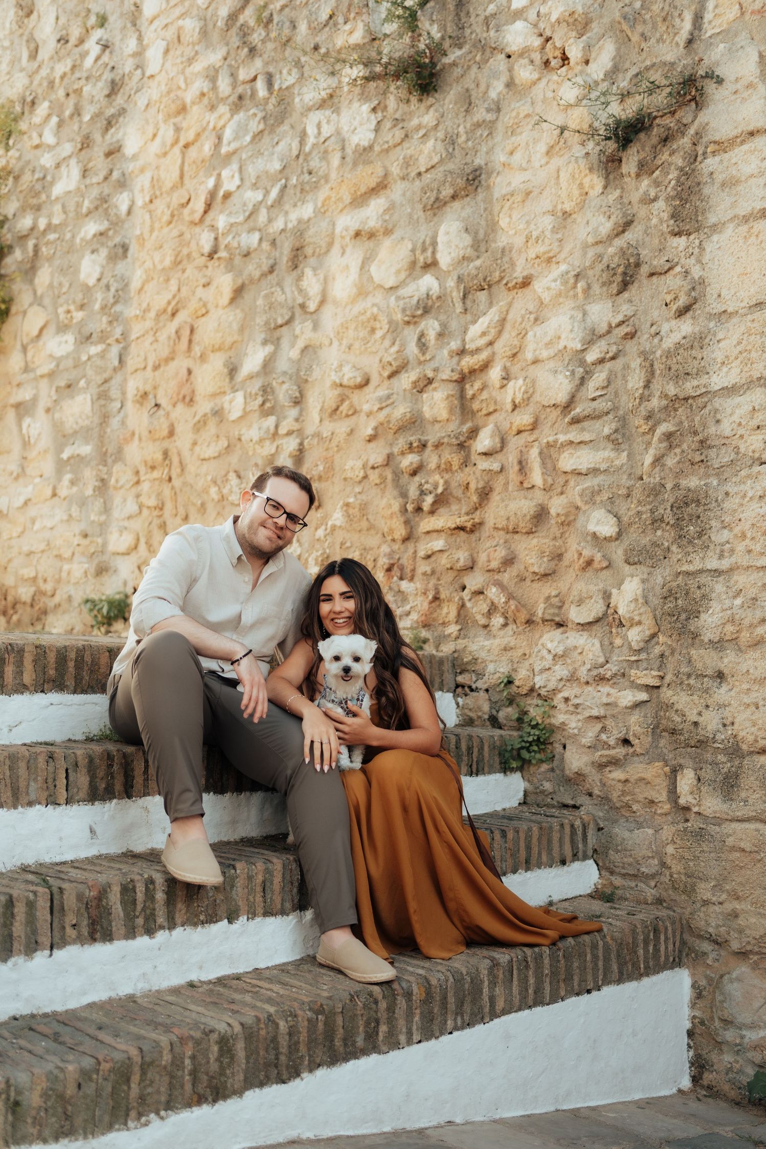 Couple sitting on stone steps with small white dog, woman in rust dress, man in beige shirt, against rustic stone wall.