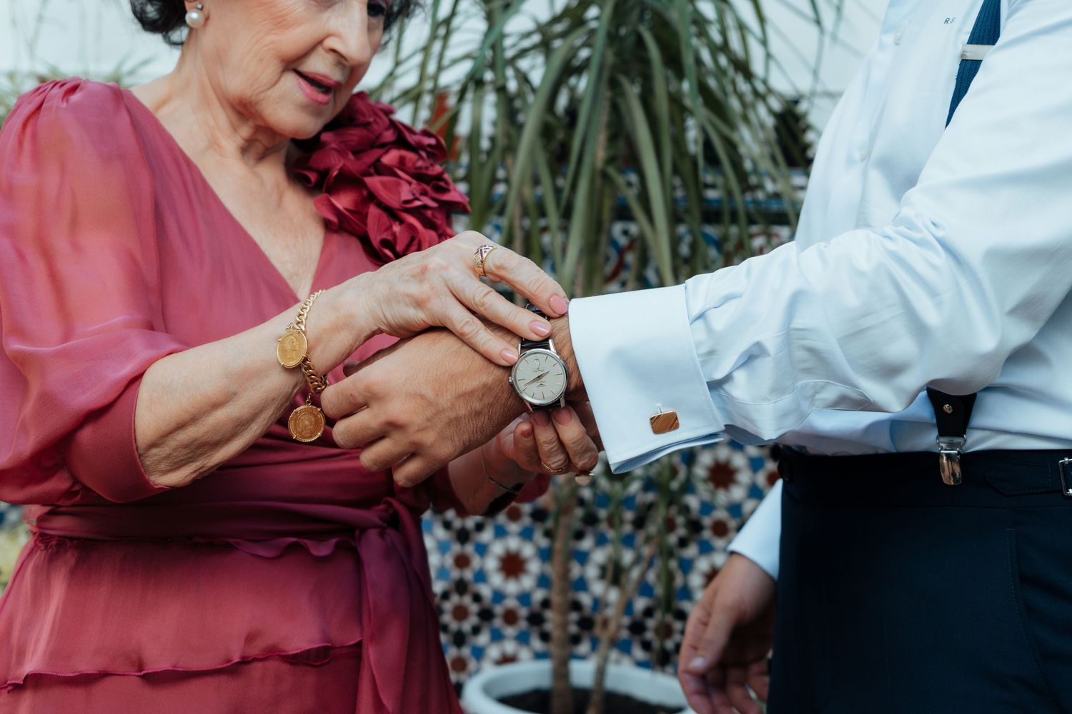Elderly woman in red dress helping groom adjust watch cufflink on white shirt outdoors.