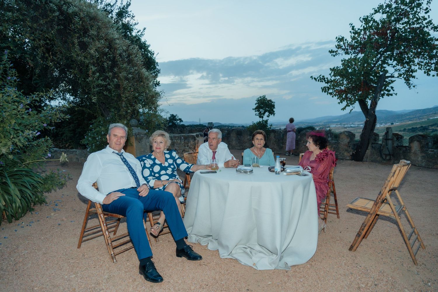 Five wedding guests seated around a table outdoors at dusk with scenic countryside views.
