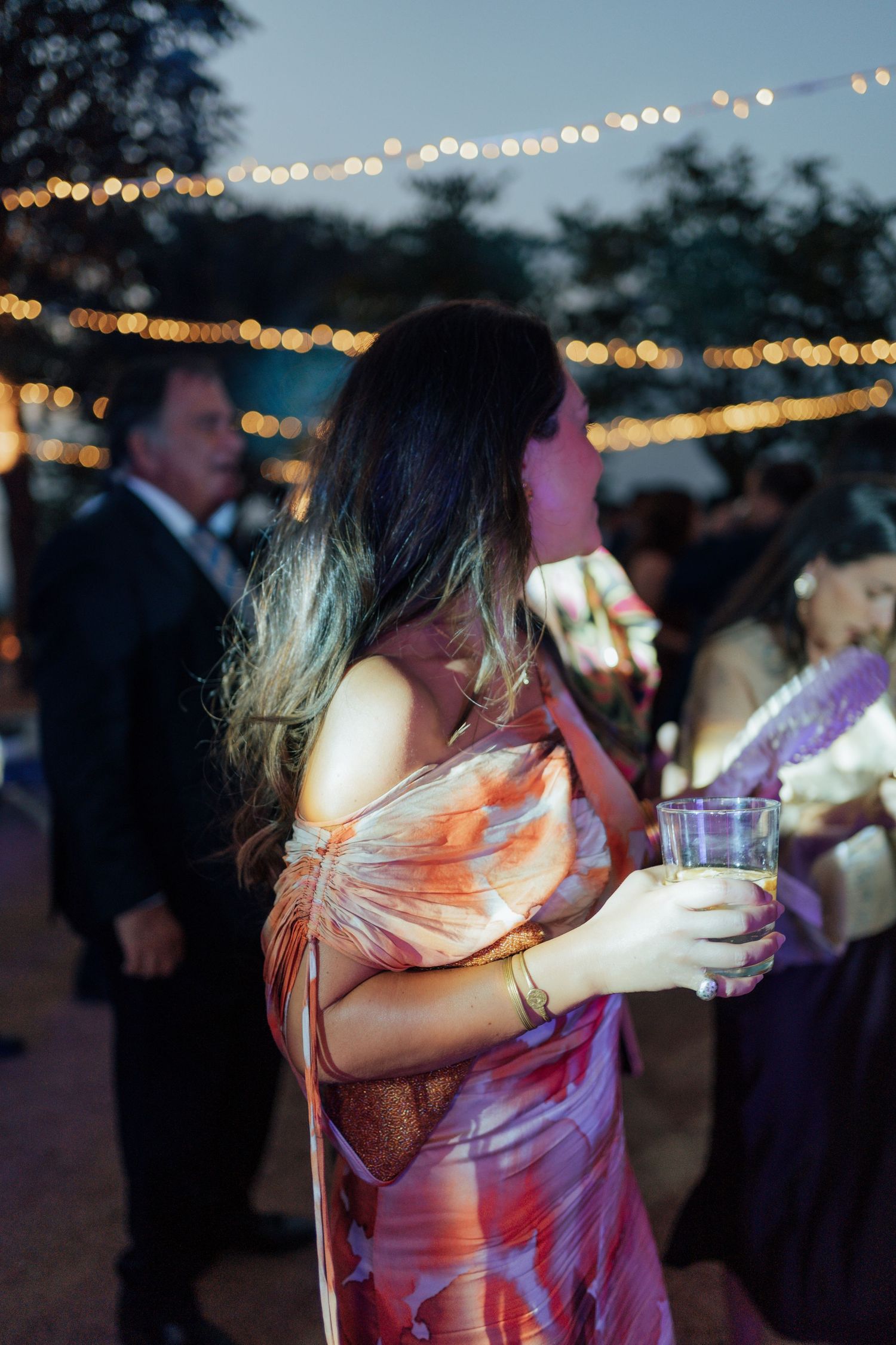 Wedding guest in floral off-shoulder dress holds drink at elegant outdoor evening reception.