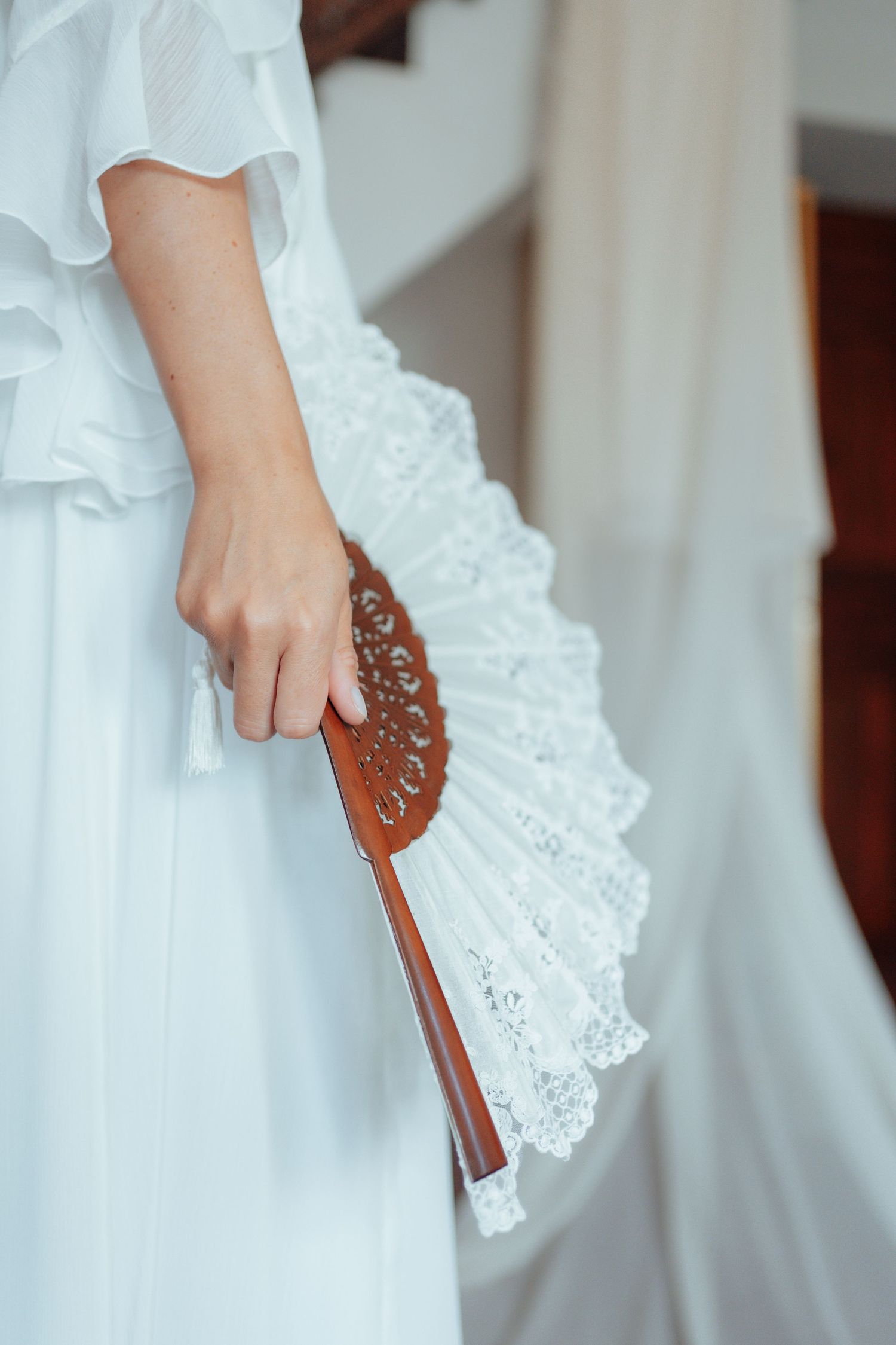 Bride holding ornate wooden and lace hand fan in white wedding dress close-up.