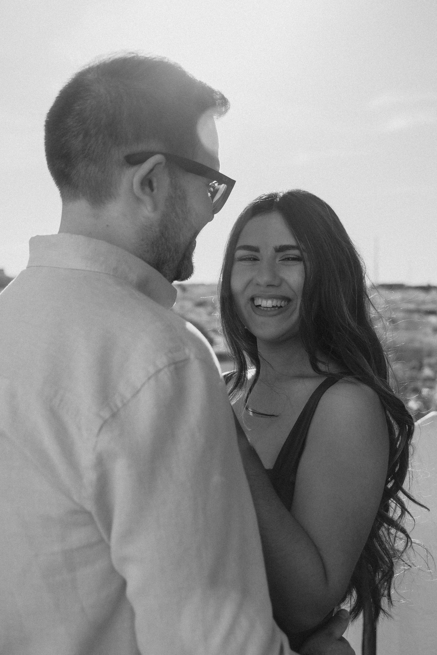 Black and white photo of a smiling woman and a man with glasses sharing a joyful moment outdoors.