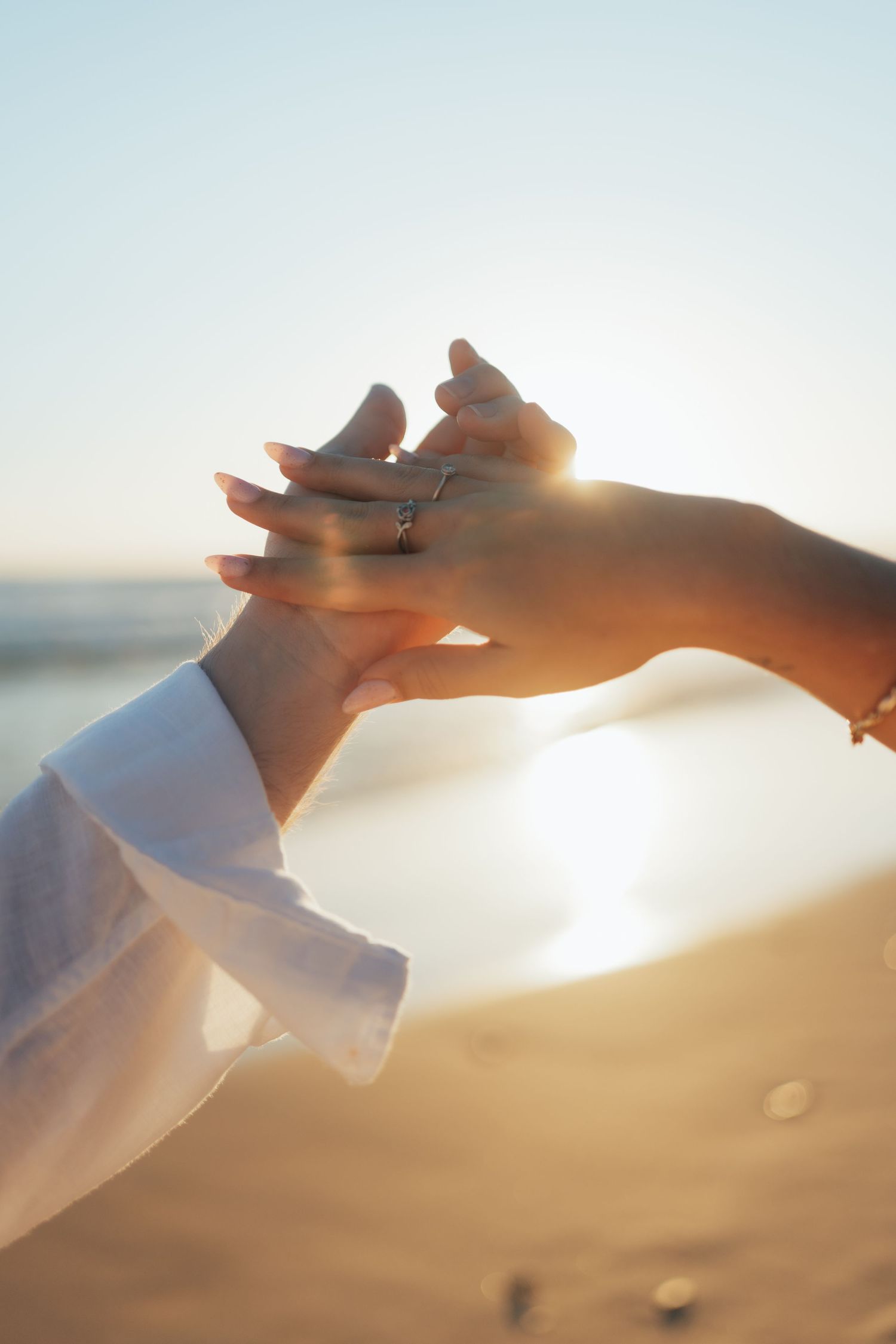 Two hands touching at sunset on a beach, with a diamond engagement ring sparkling in the golden light.