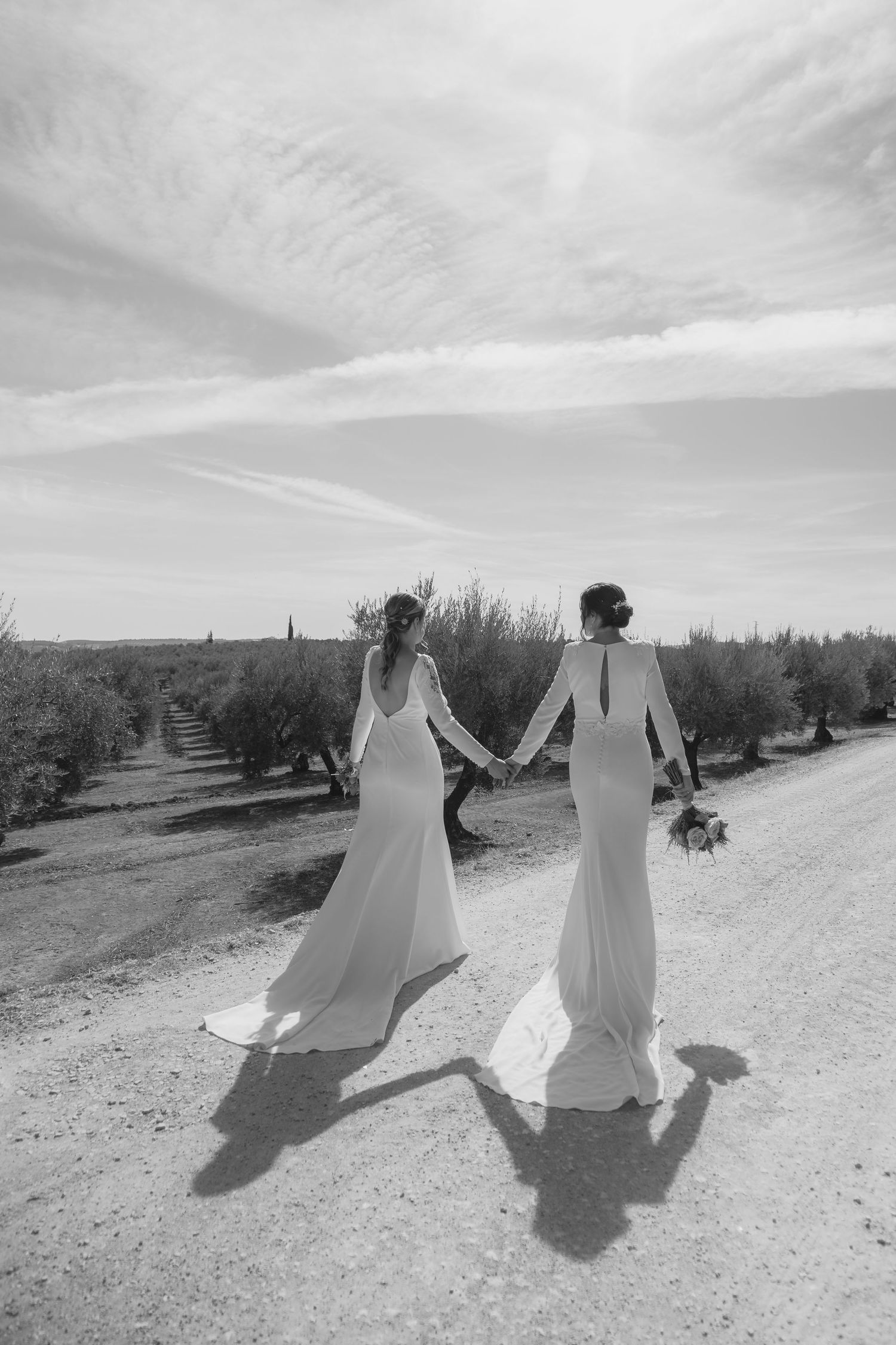 Two brides in elegant white long-sleeve gowns hold hands walking down a sunny dirt road in black and white.