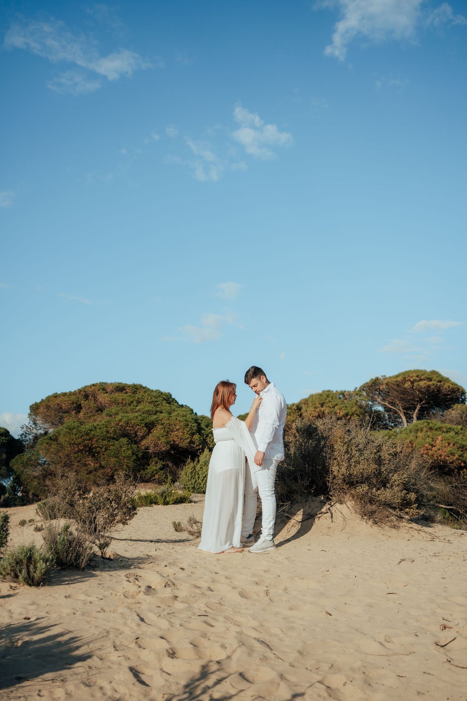 Couple in white outfits holding hands on sandy beach dunes with pine trees and blue sky backdrop.