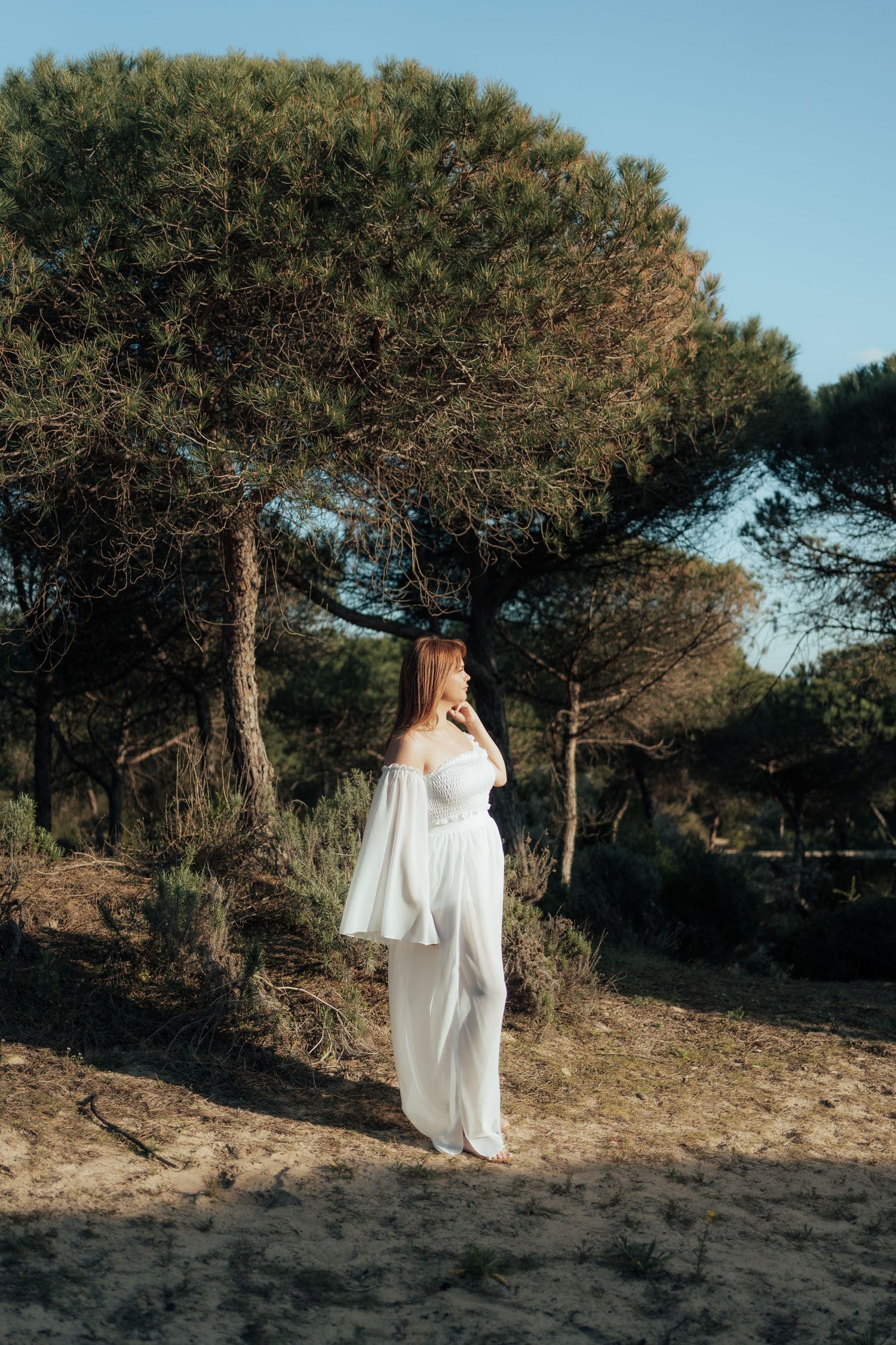 Woman in white off-shoulder outfit standing on sandy ground surrounded by pine trees in soft natural light.