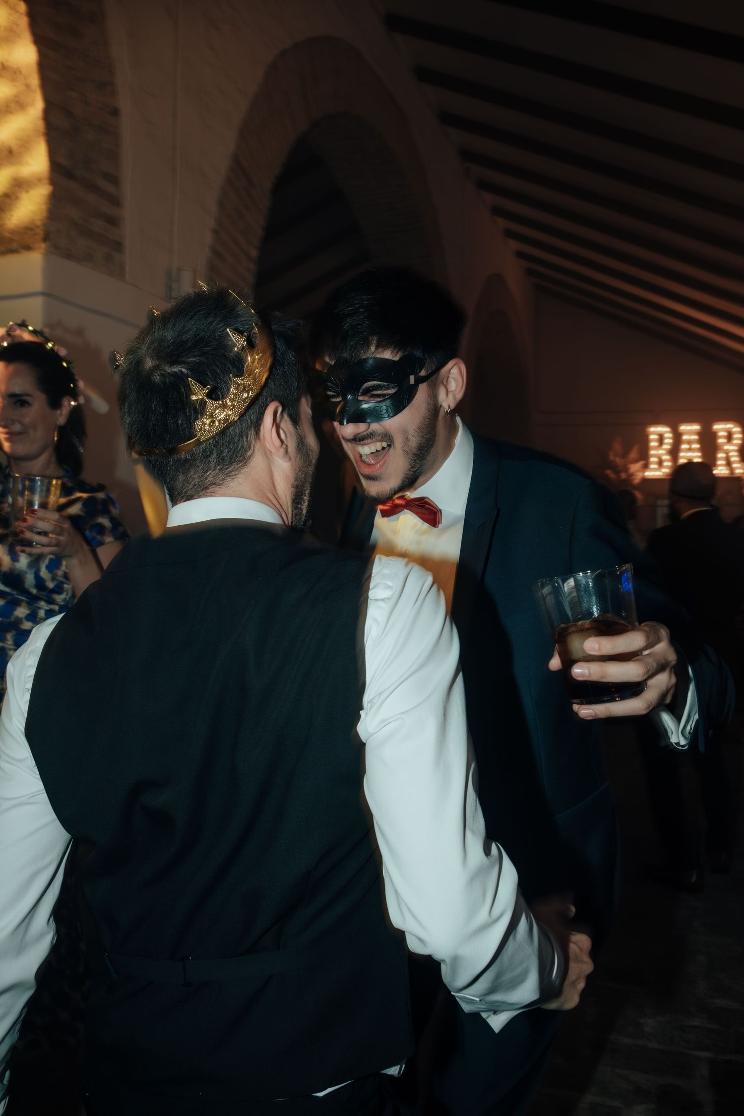 Two men wearing masquerade masks socialize and laugh together at an elegant masquerade ball event.