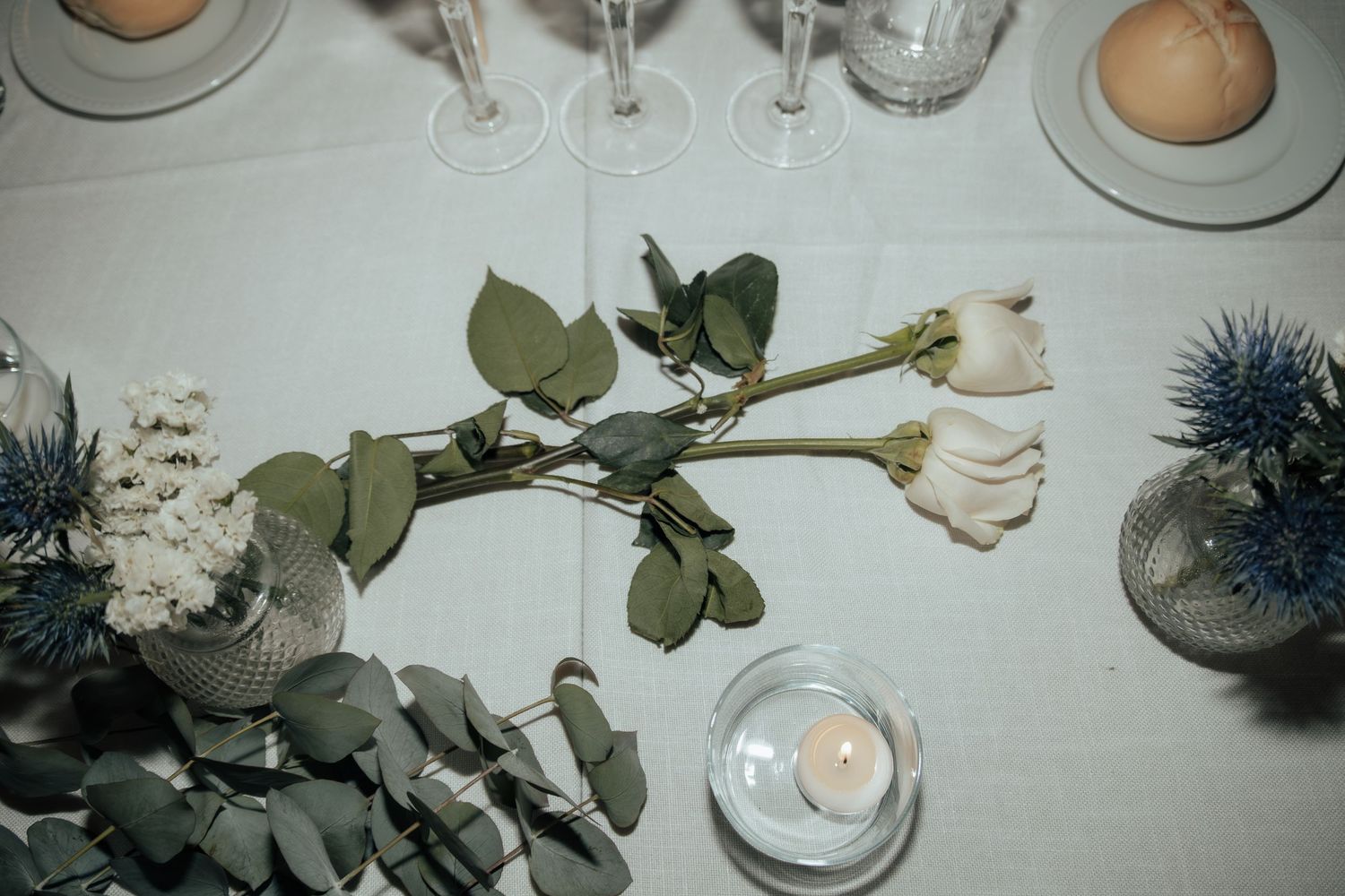 Elegant table centerpiece with white roses, eucalyptus, blue thistles, and candles at a wedding.