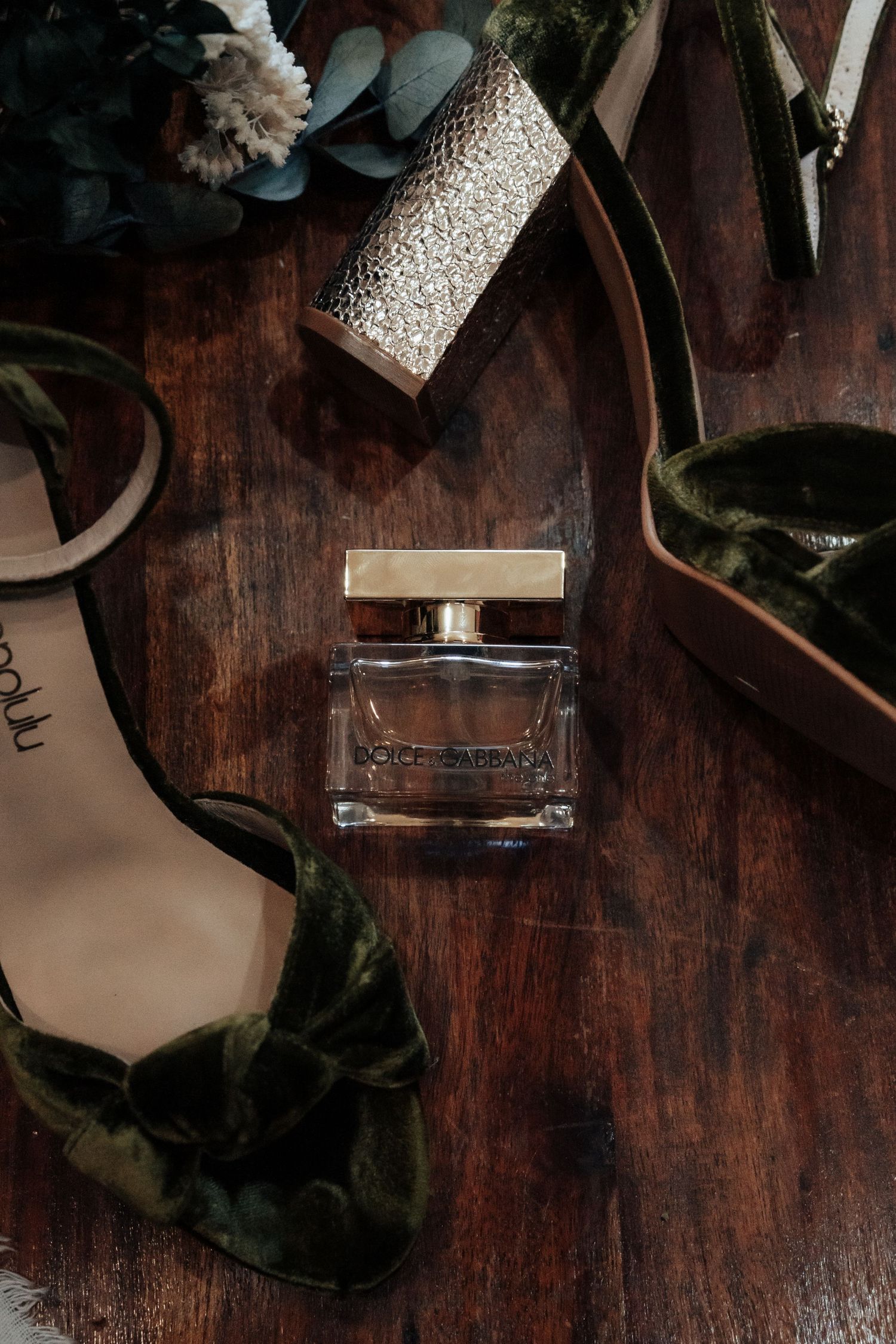 Dolce and Gabbana perfume surrounded by green velvet heels and gold bracelet on wooden surface.