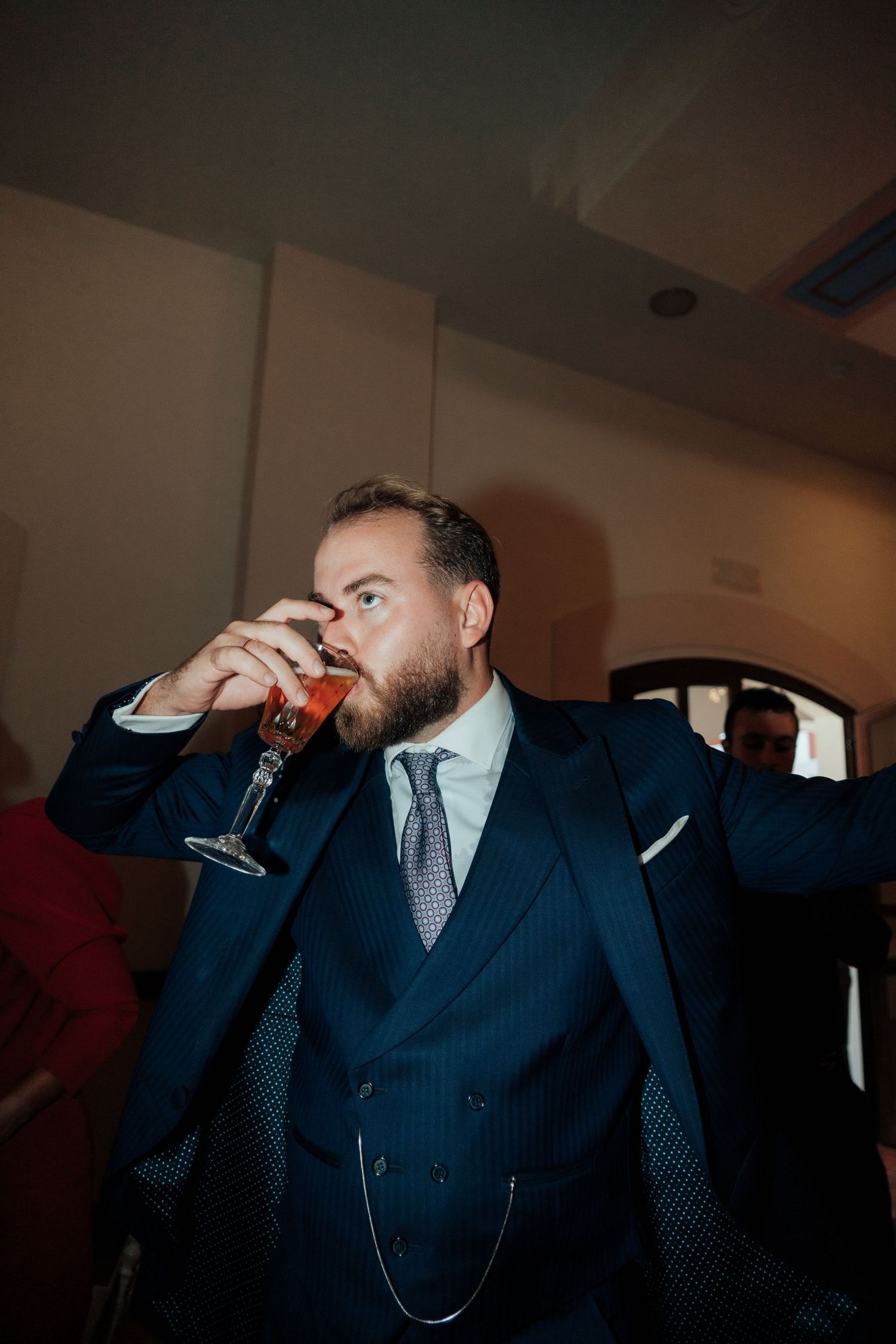 Bearded man in navy double-breasted suit drinks red cocktail from champagne flute at wedding party.