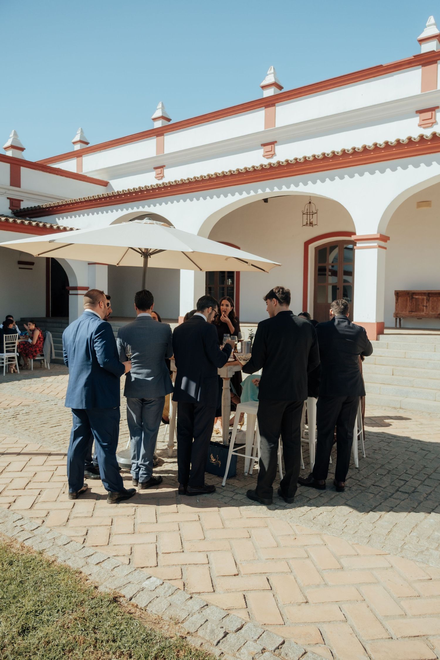 Wedding guests gathered under white umbrellas at outdoor bar in sunny courtyard.