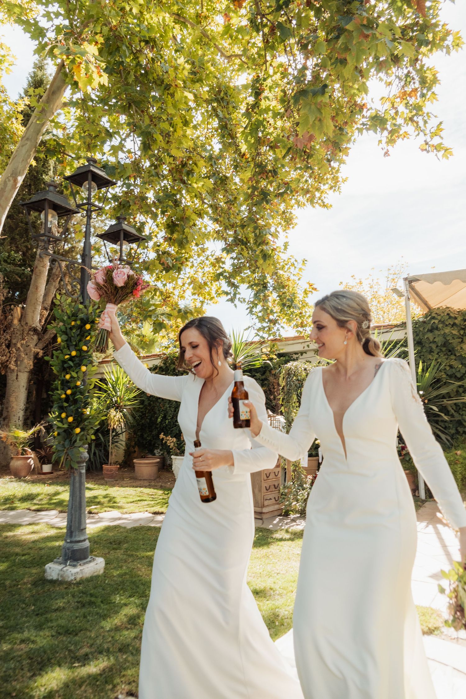 Two brides in white gowns celebrating outdoors, holding bouquets and beer bottles in a garden.